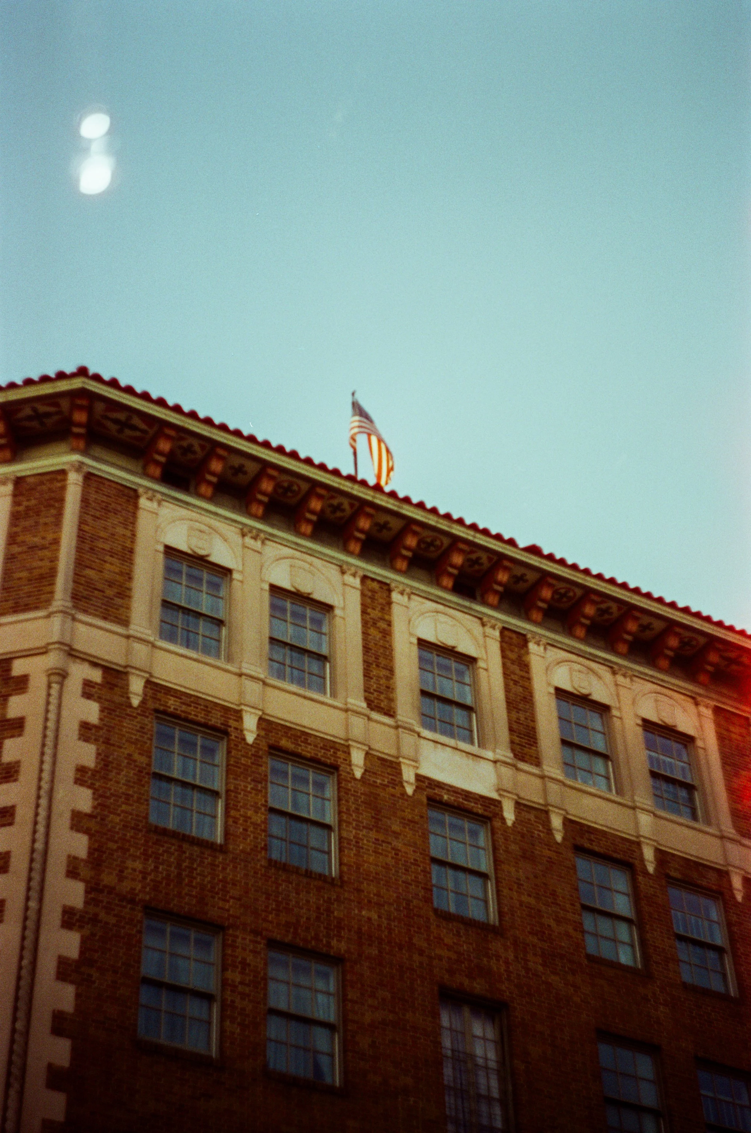 A multi-story brick building with white decorative trim, several windows, and an American flag on the roof against a light blue sky.