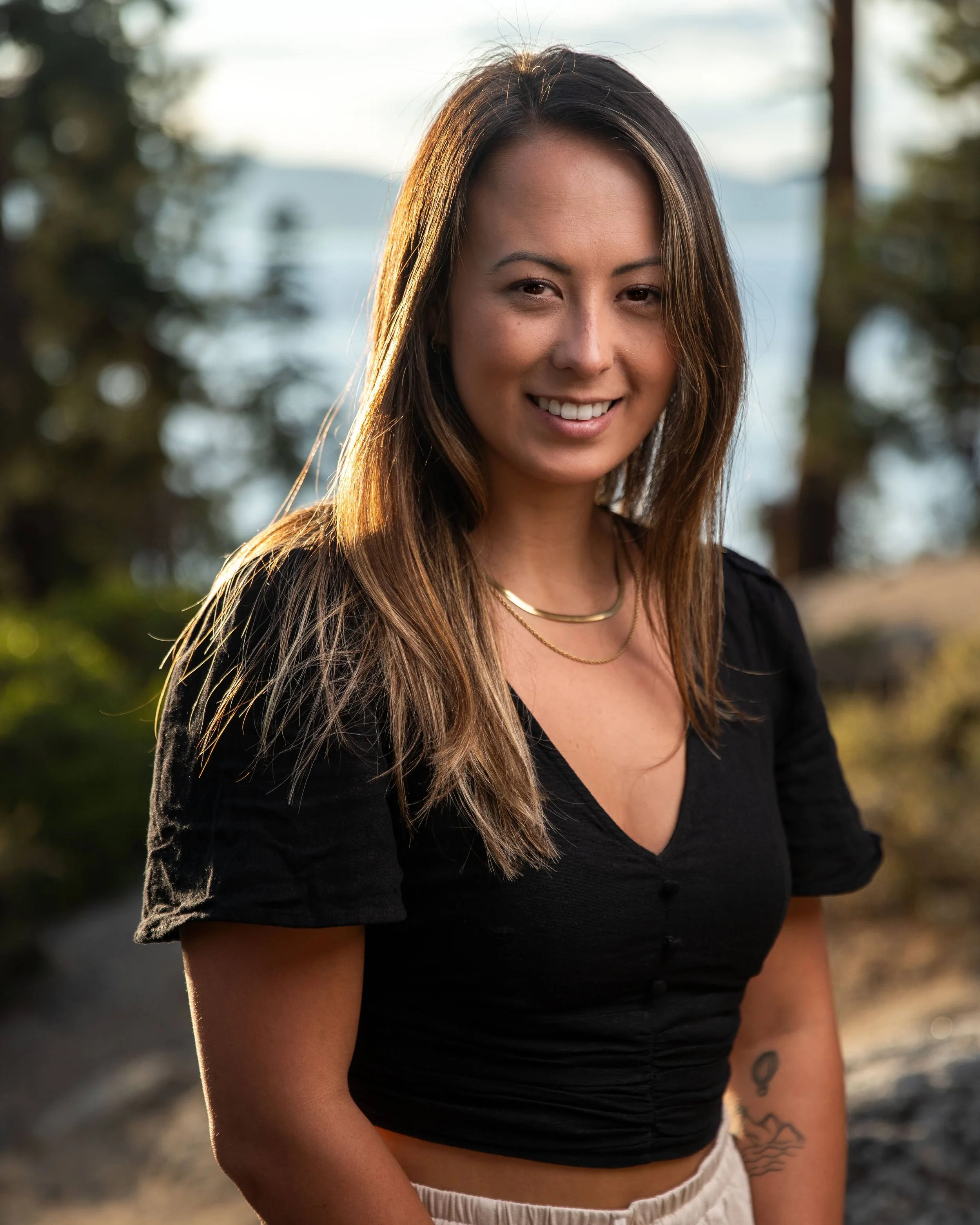 A woman with long brown hair smiling outdoors in front of trees and a body of water, wearing a black top and layered necklaces.