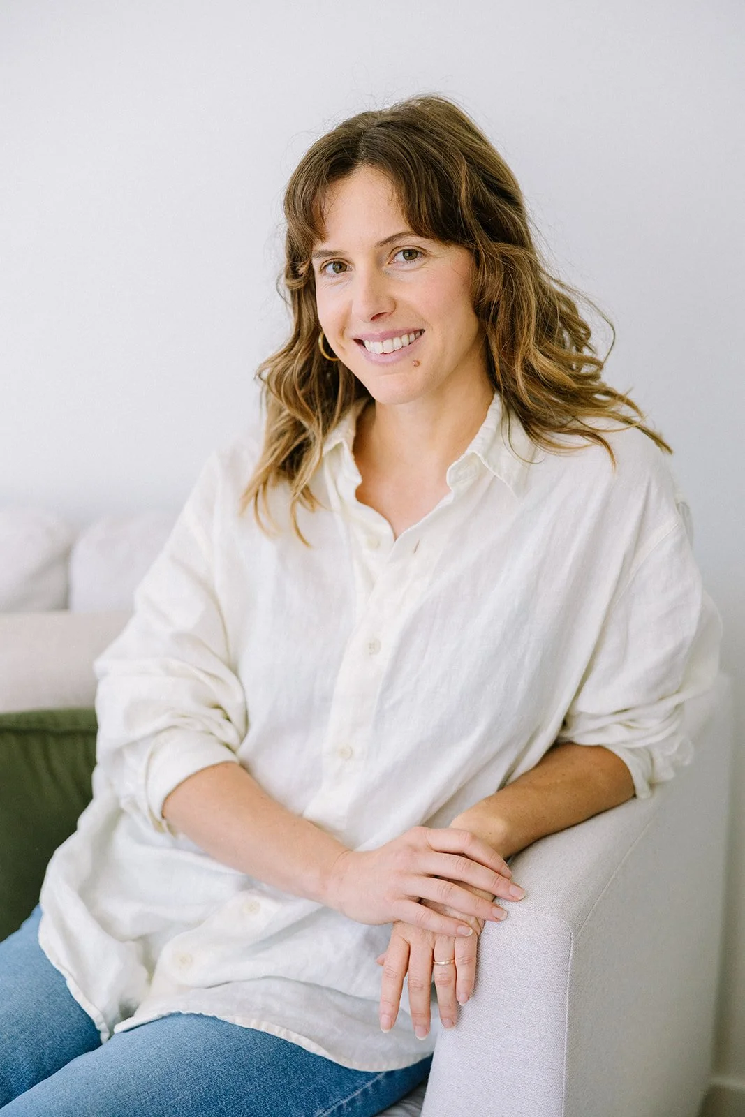 A woman with shoulder-length wavy brown hair wearing a cream-colored button-up shirt and blue jeans, sitting on a light-colored armchair with a white background and a couch with a green pillow behind her.