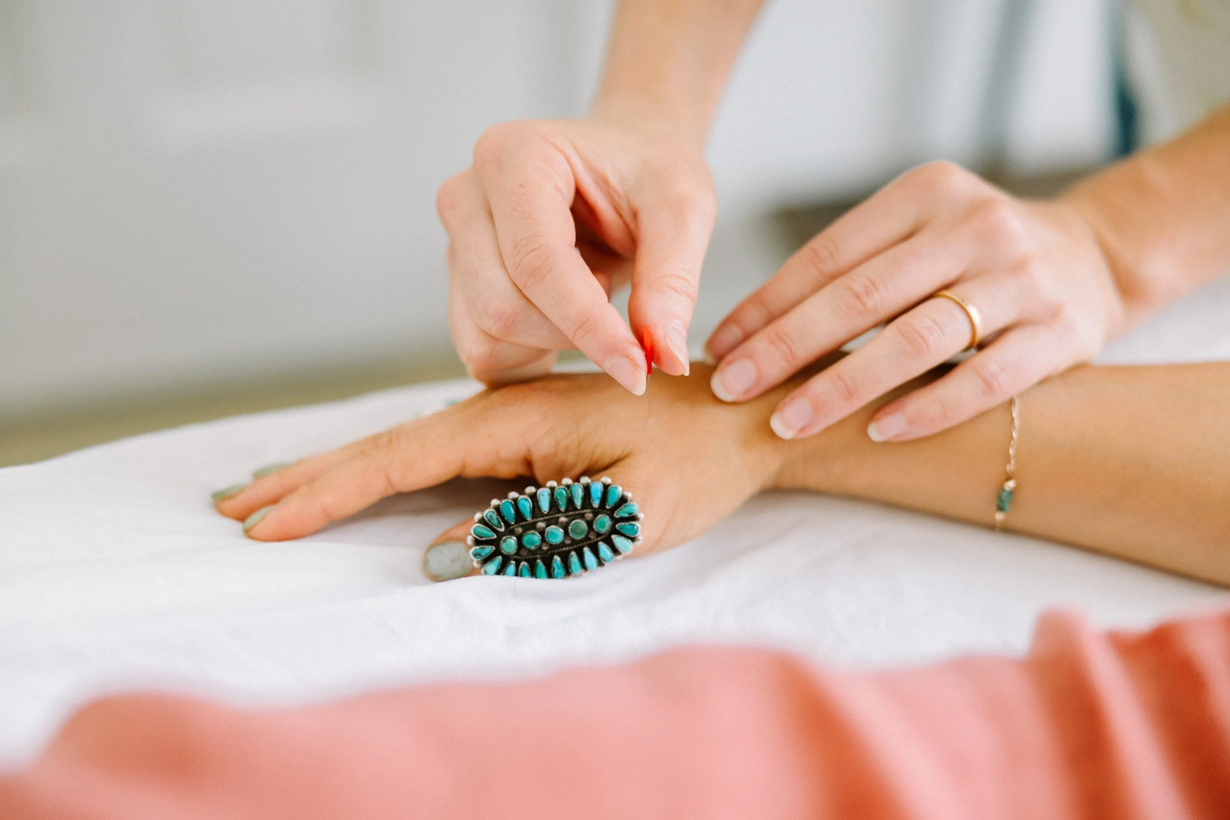 Person receiving a manicure with turquoise nail polish, wearing a ring and bracelet, with a beauty or healthcare professional applying nail polish or finishing touches.