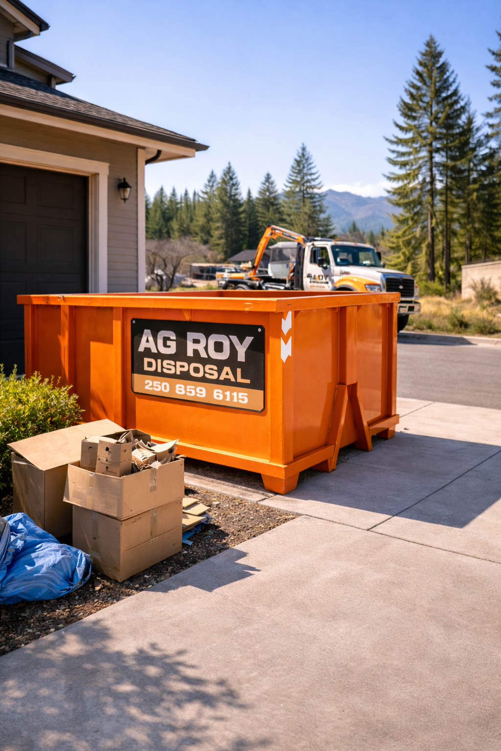 Orange dumpster labeled AG ROY DISPOSAL on a driveway with cardboard boxes and trash bags nearby; a white and orange pickup truck with a crane in the background, surrounded by trees and mountains.