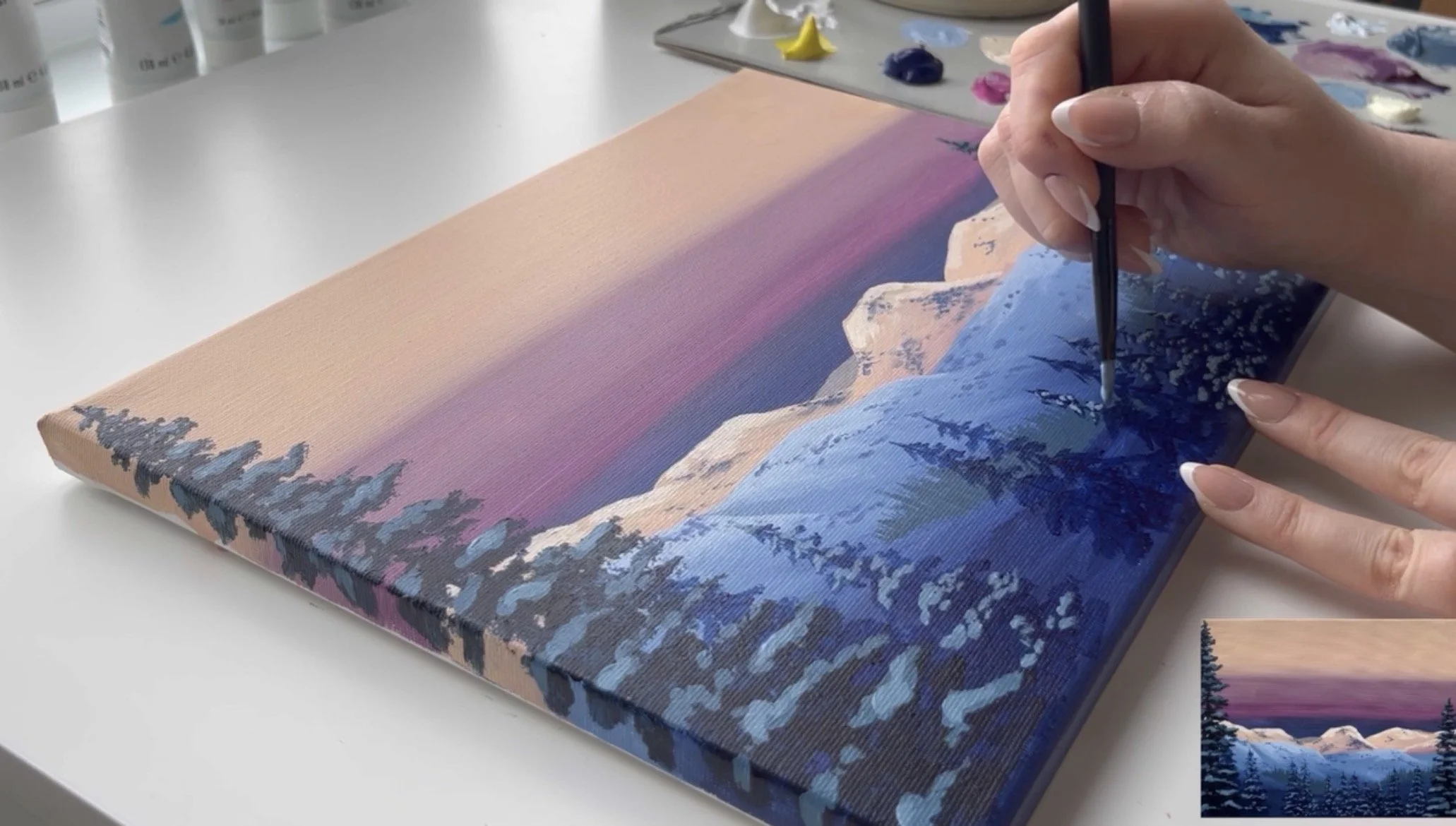 An artist's hands painting a realistic acrylic landscape painting of a mountain range at sunset with vibrant sky. It is a still image from a video tutorial the Landscape Masterpiece Class.