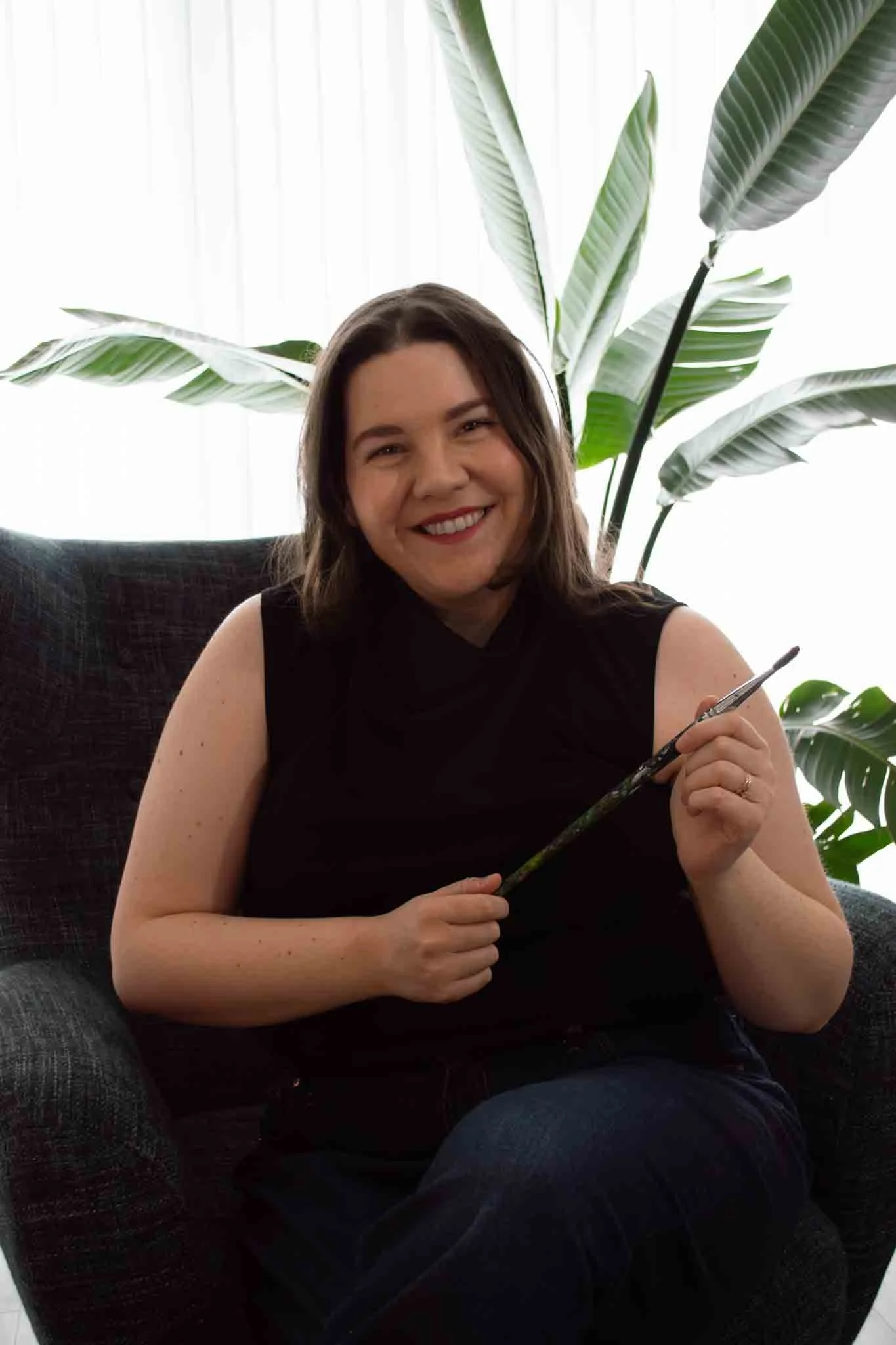 Professional acrylic landscape artist portrait featuring a woman holding a paintbrush in a bright, modern studio setting with lush greenery.