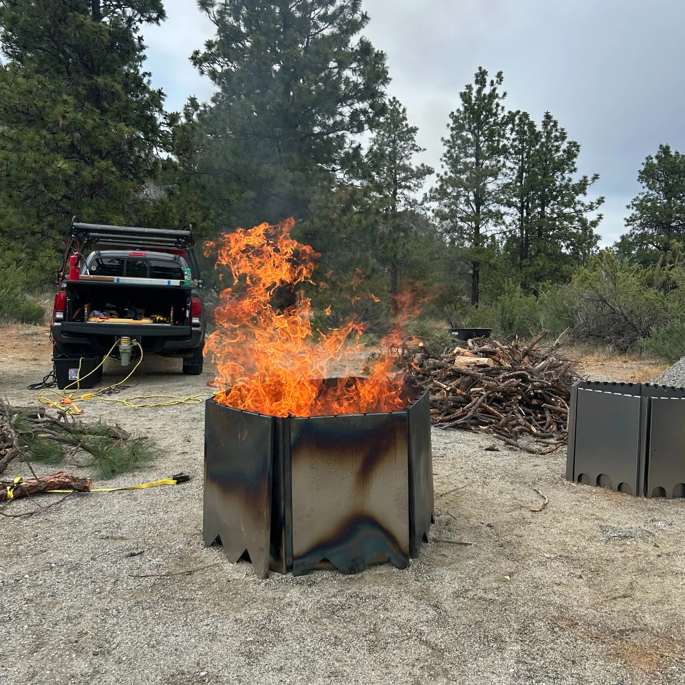 A controlled outdoor burn in a biochar kiln made of steel metal, with fire and flames, surrounded by wood piles and a truck with firefighting equipment in a forested area.