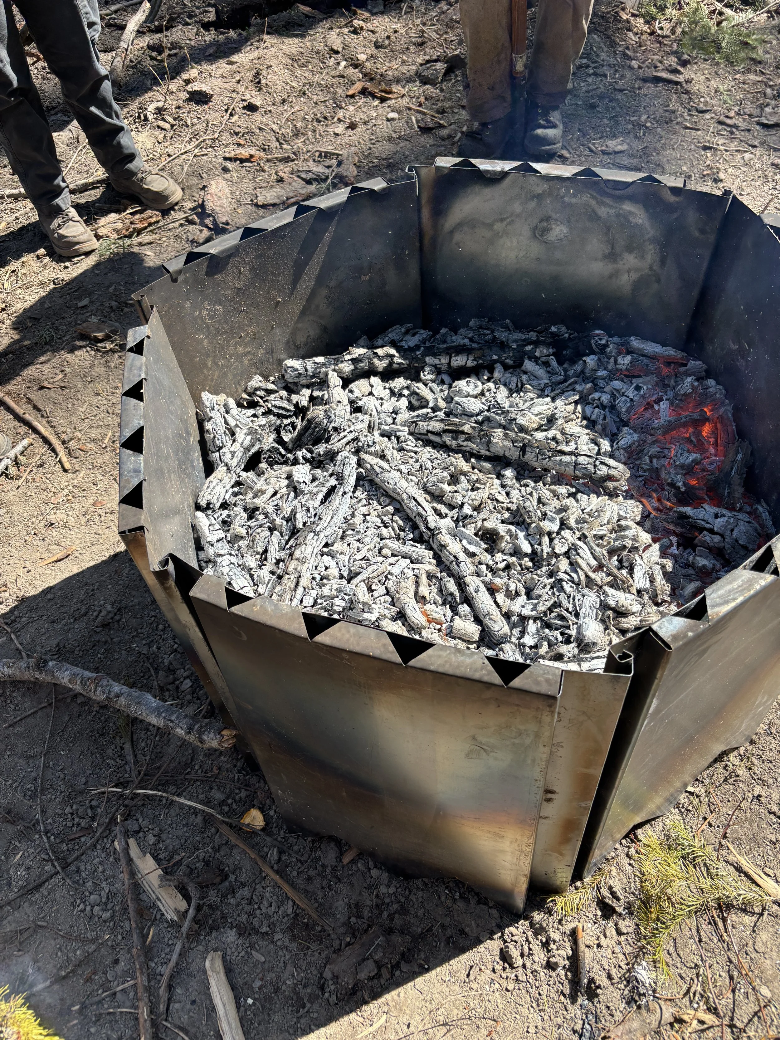 A biochar kiln near the end of a burn, right before quenching, where the char has a bit of ash on top, with small flames burning on one side, outdoors on dirt ground surrounded by tree debris.