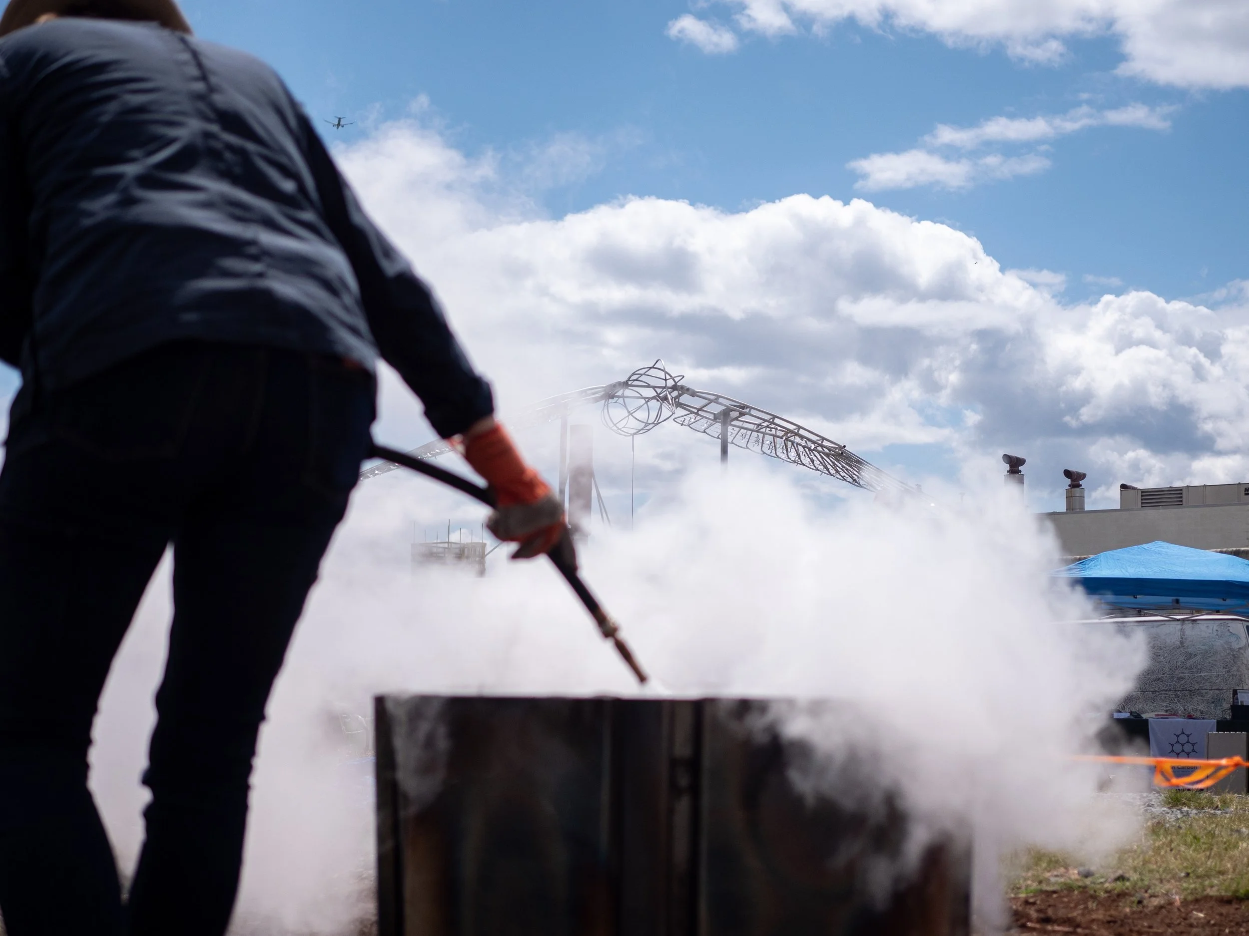 Person using a hose to quench the biochar, emitting steam, outdoors with cloudy skies.