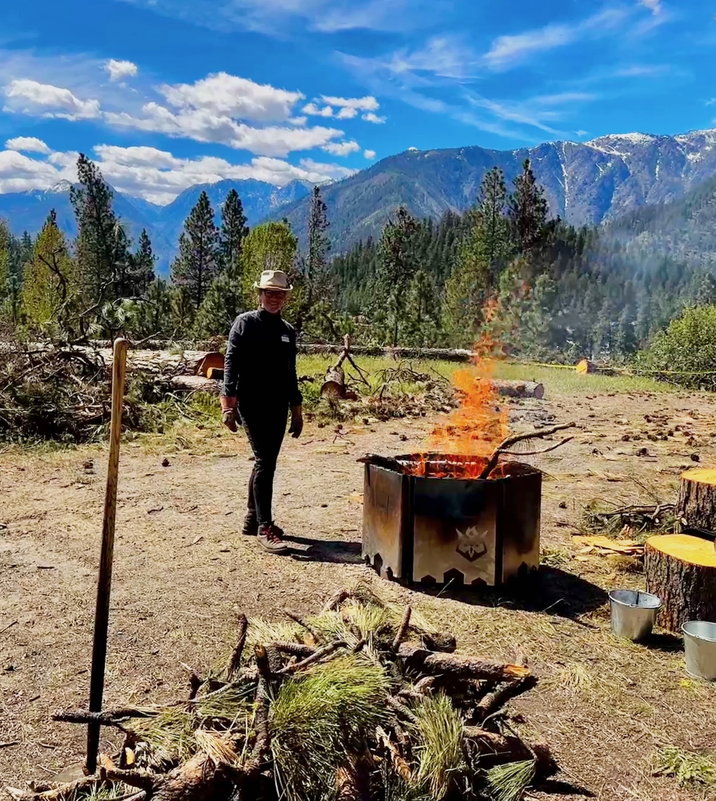 Person in outdoor gear standing next to a small controlled fire in a biochar kiln the size of a fire pit, with a forested hillside and blue sky in the background.
