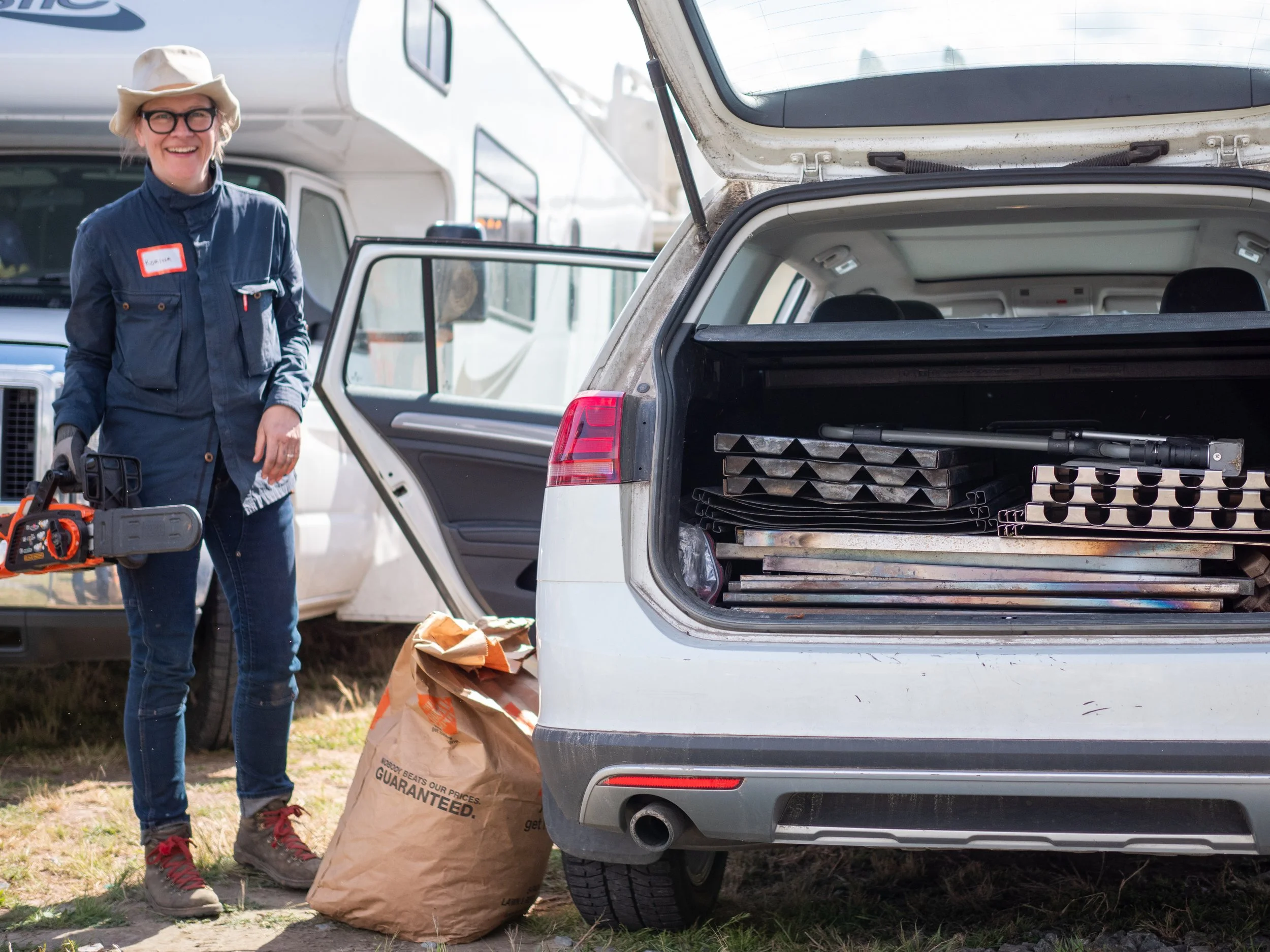 A woman with glasses and a hat standing outdoors next to an open car trunk filled with metal tools and biochar equipment, holding a power tool in her right hand, with an RV in the background and a paper bag on the ground.