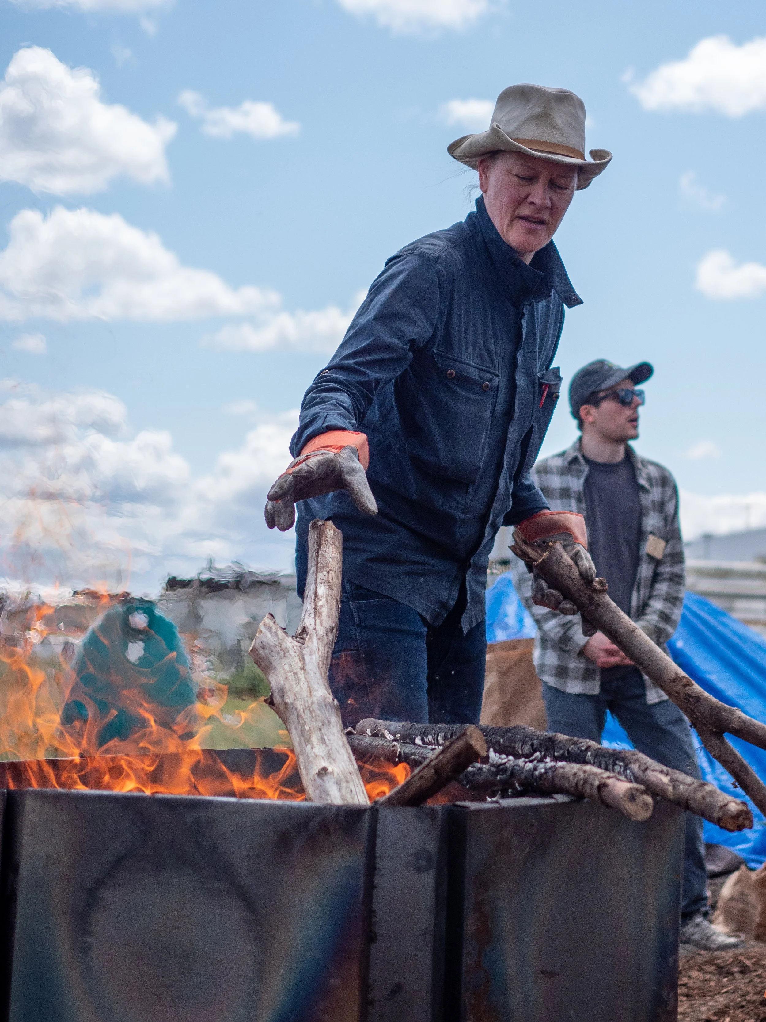 A woman wearing a beige hat, blue jacket, and gloves tending the flame of a biochar kiln with logs outdoors under a blue sky with clouds, with a young man in sunglasses and a plaid shirt in the background.