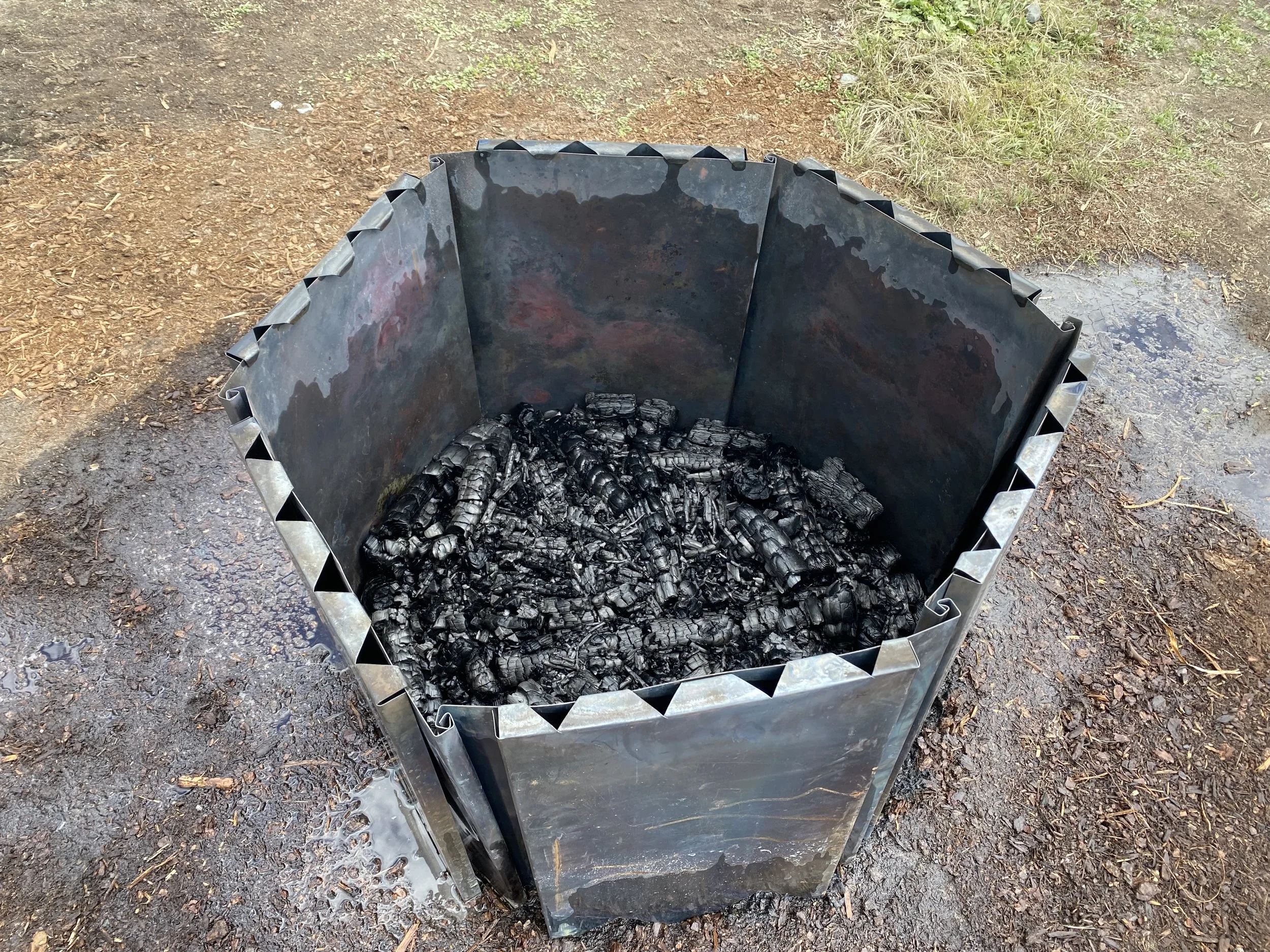 A biochar kiln filled with fresh charred wood or charcoal on an outdoor ground surface.