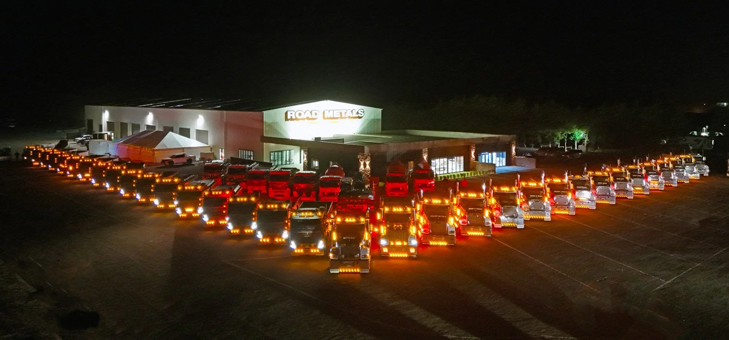 A large fleet of trucks at night with their lights on in front of a large workshop and office complex