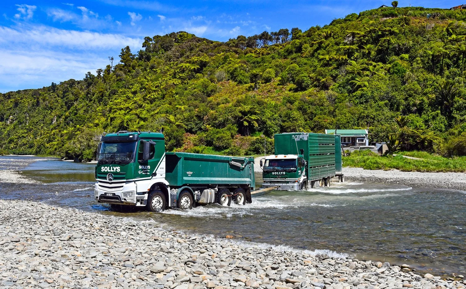 two green and white trucks crossing a river, the front tip truck is towing the back stock truck
