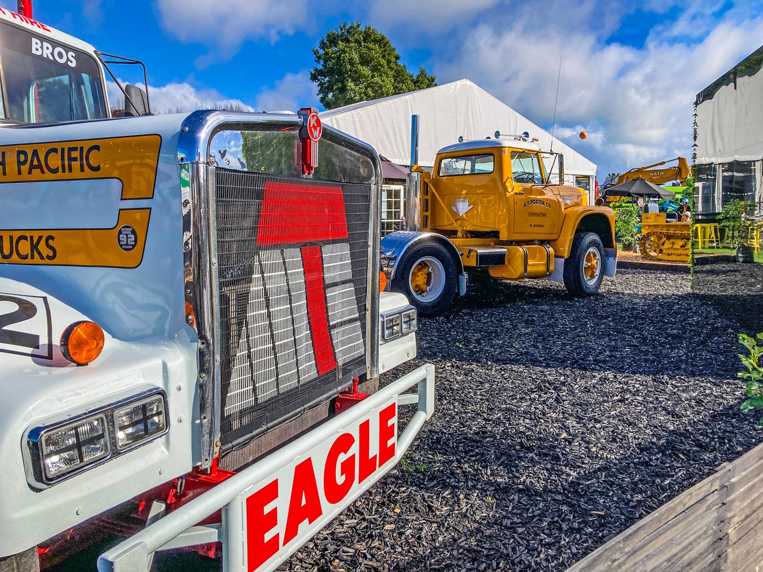 The bonnet and grill of a white truck with a yellow truck in the background, both are sitting on bark with a marque in the background at a show