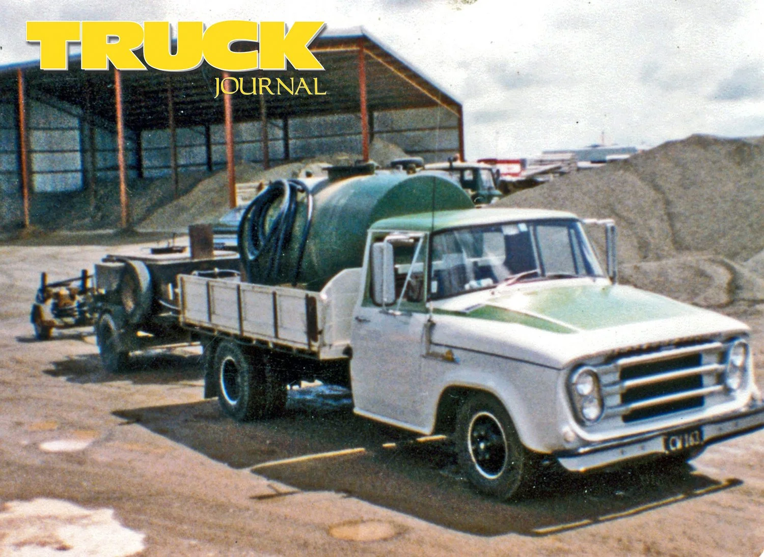 A white and green light truck with a tank on its deck and two trailers in a contractors yard with gravel piles and sheds in the background