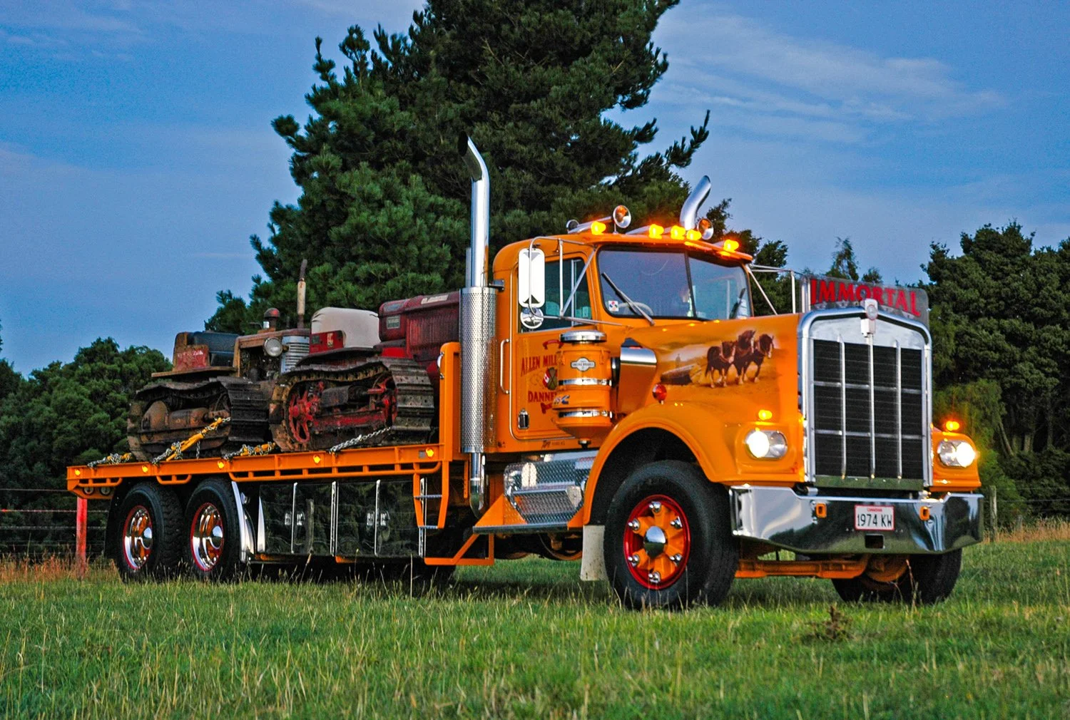 A yellow flat deck truck with 2 bulldozers on the deck parked on grass