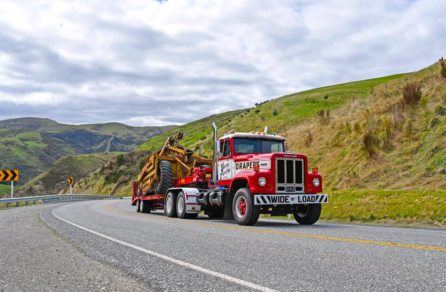 A red and white truck with a low loader trailer with a scraper bowl  climbing a hill road with hills in the background