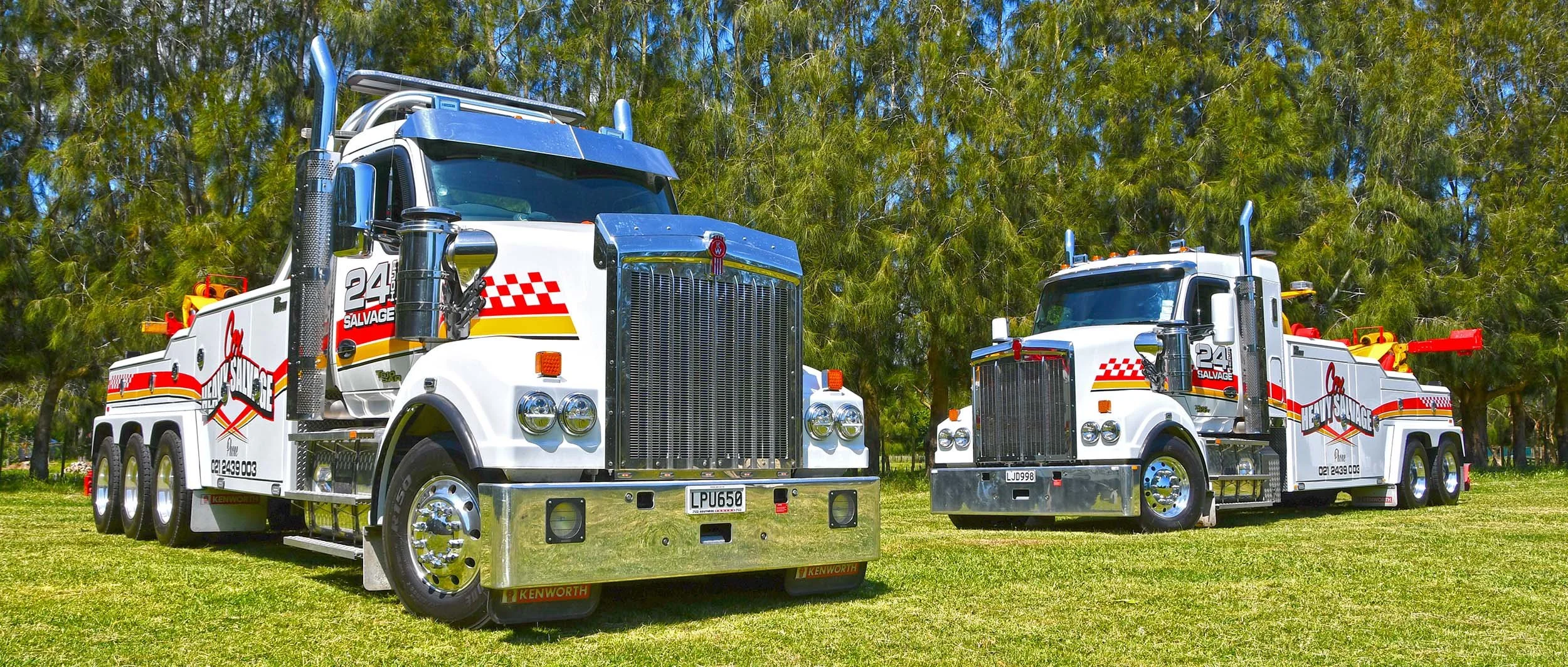 Two white tow trucks with red and yellow stripes parked on a grassy area beneath trees.