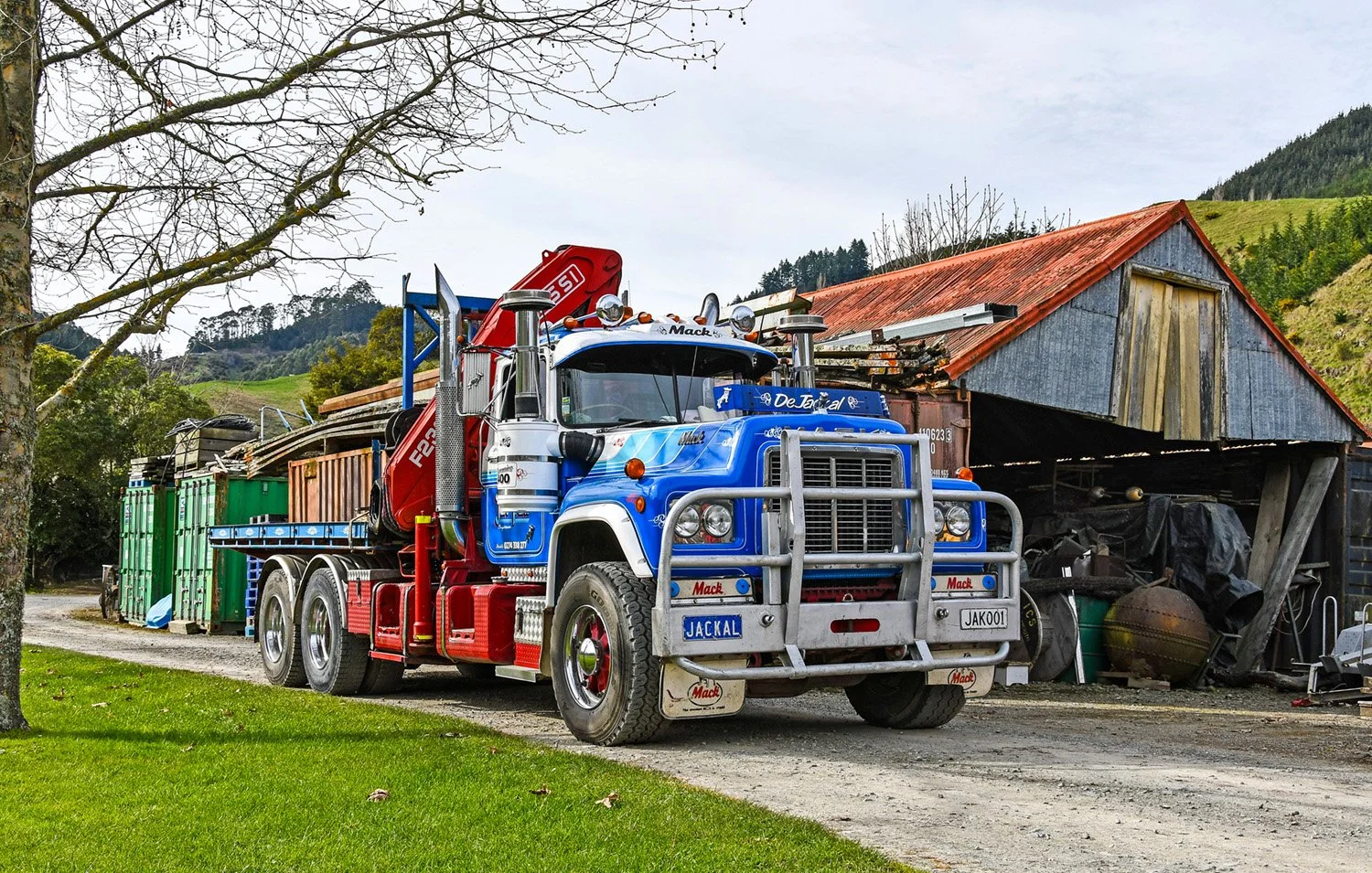 A white, red and blue truck sitting between a terr and a garage. The truck has a flat deck and crane.