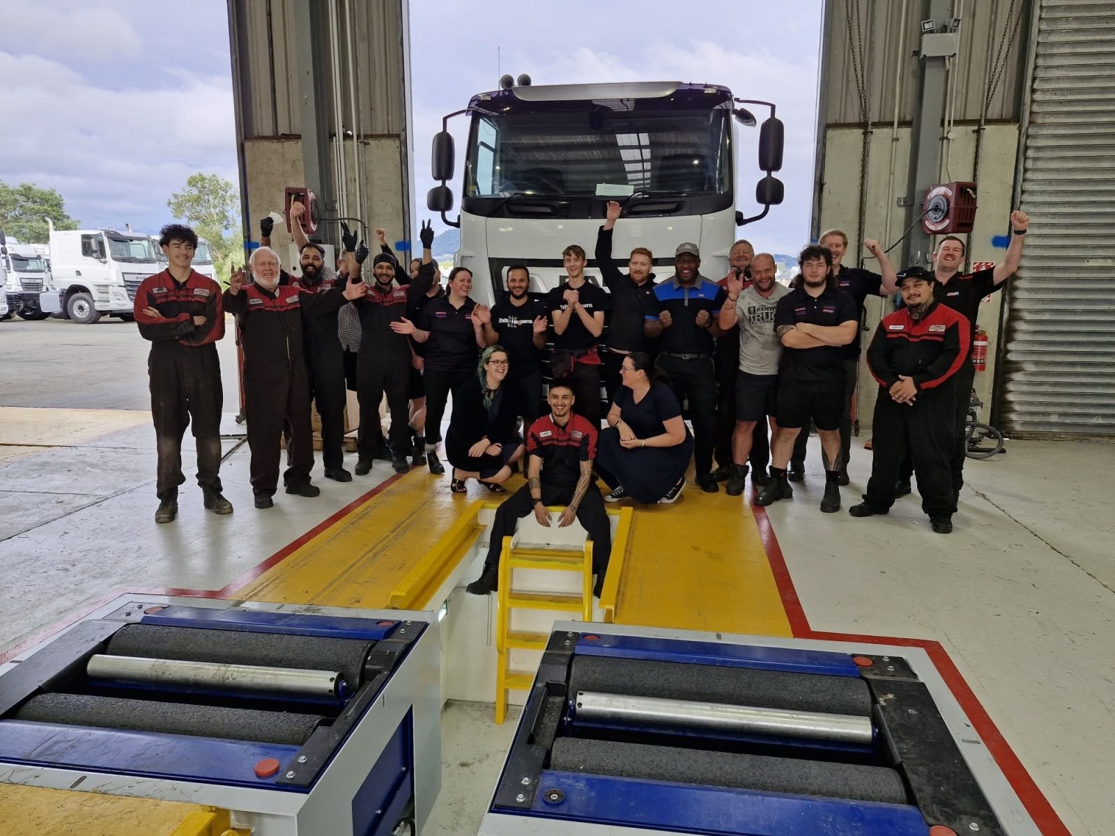 A group of mechanics stand at the end of a workshop pit with a truck and an open door behind them