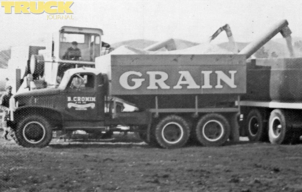 A truck with a v bin is both being loaded from farm machinery and also loading a tip truck parked behind it