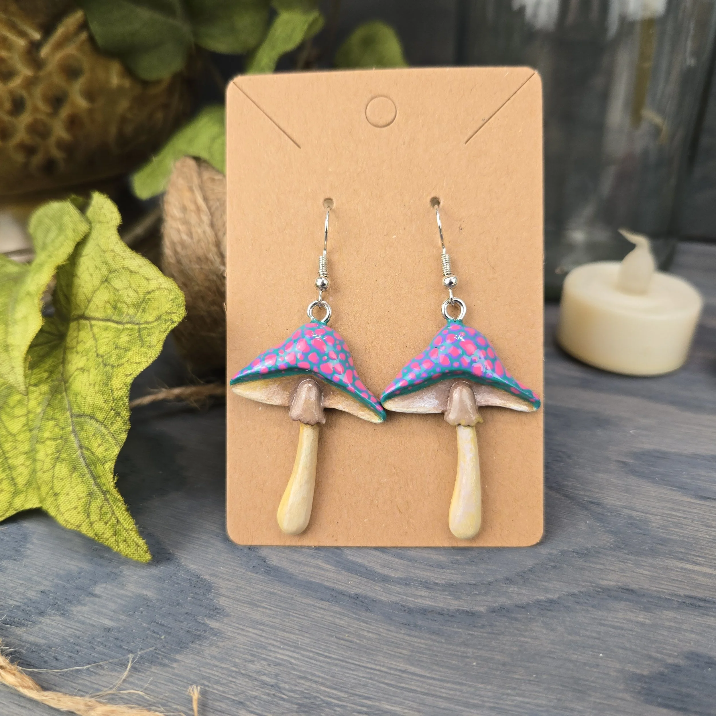 Colorful mushroom-shaped hook earrings with pink and blue polka-dotted caps and beige stems, hanging on a brown display card.