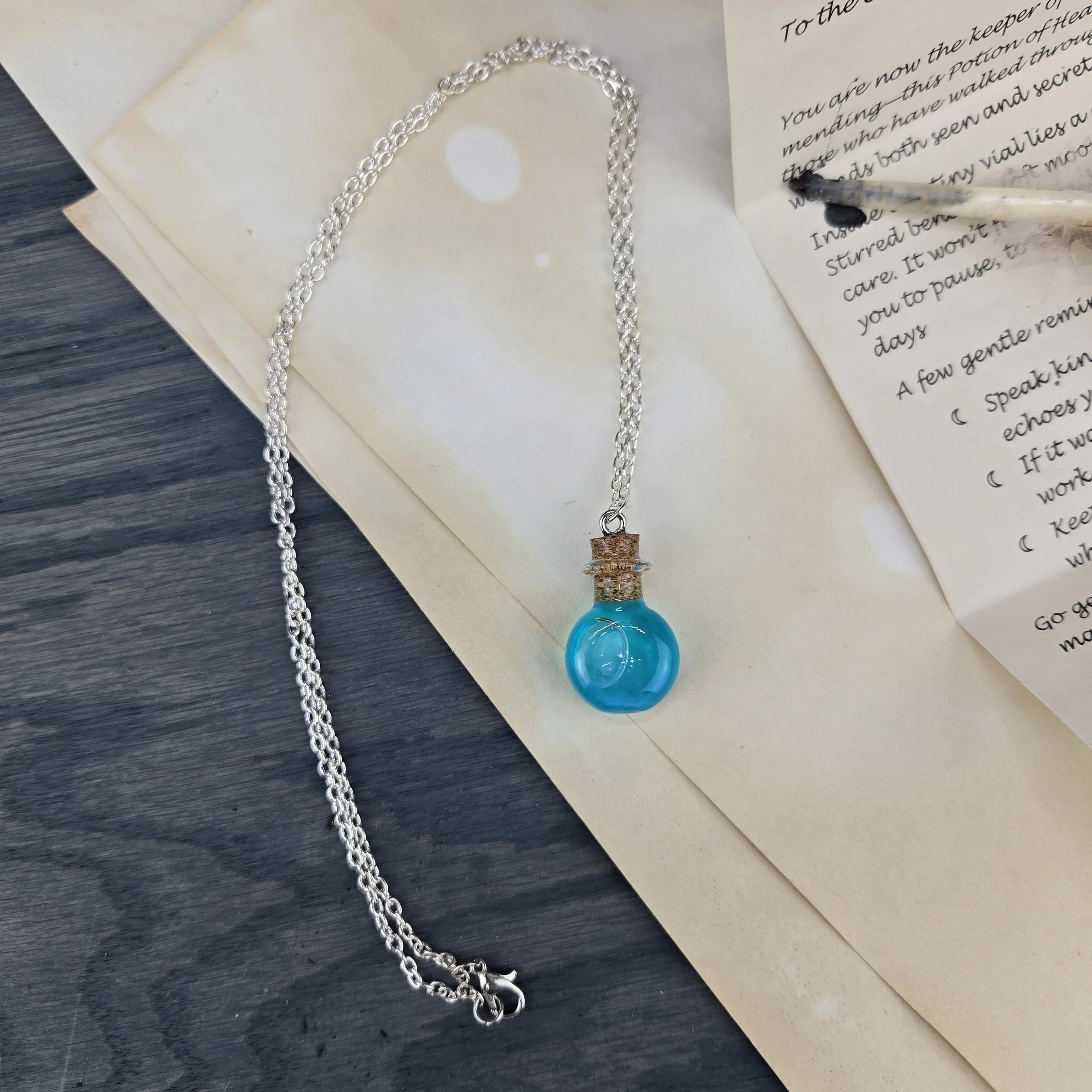 A silver chain necklace with a blue glass potion bottle pendant resting on handwritten letters on a wooden surface.