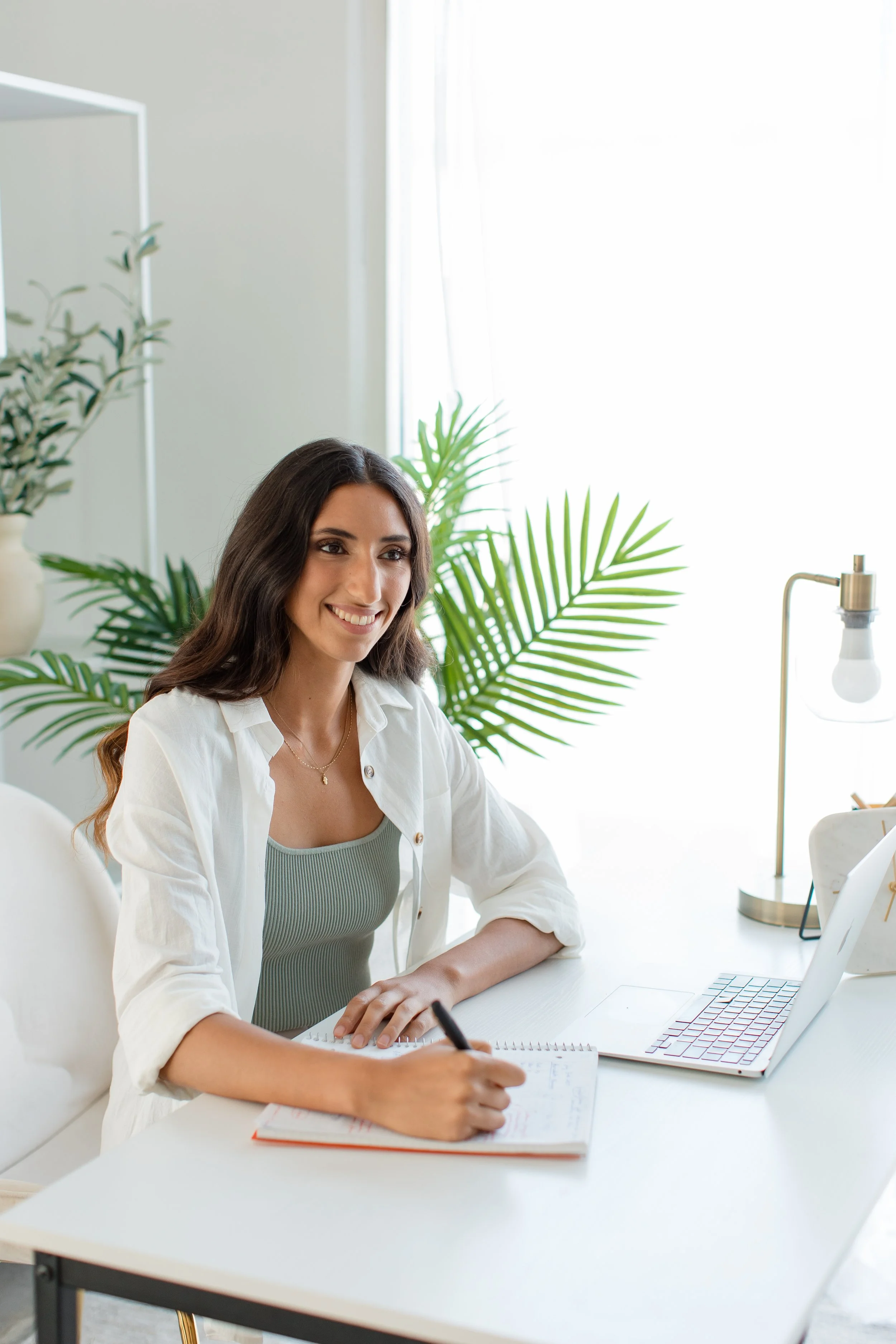 A woman with long dark hair smiling and taking notes at a white desk with a laptop, notebook, and pen, in a bright room with green plants and a lamp.