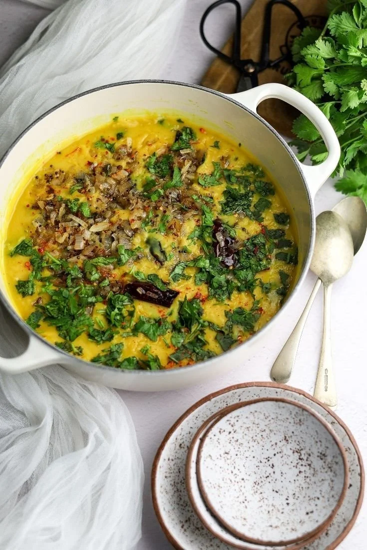 A pot of cooked yellow curry garnished with chopped herbs, dried red chilies, and onions, with a bunch of cilantro nearby and empty bowls and spoons beside it.