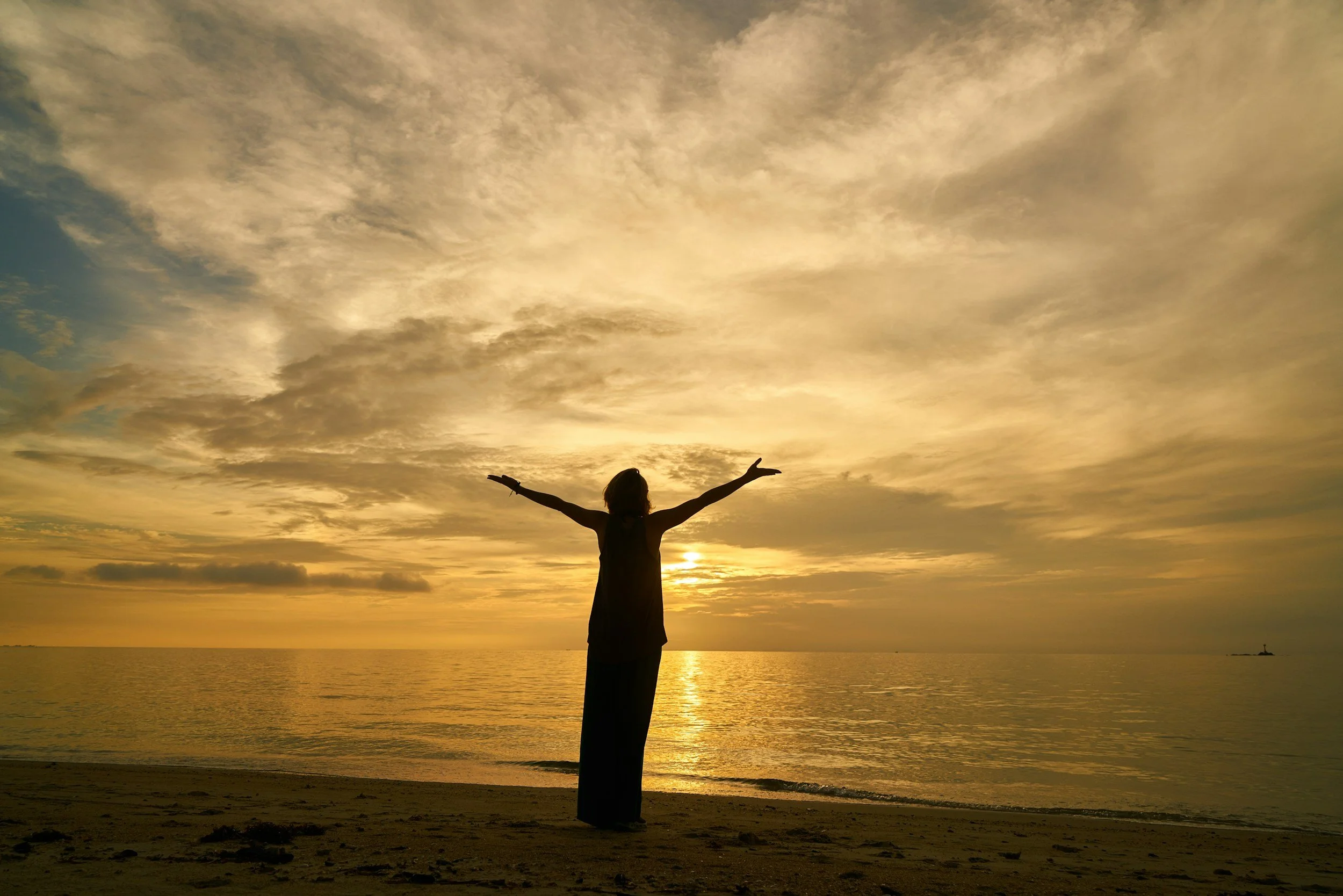 Person standing on a beach at sunset with arms outstretched, facing the ocean and sky filled with clouds.