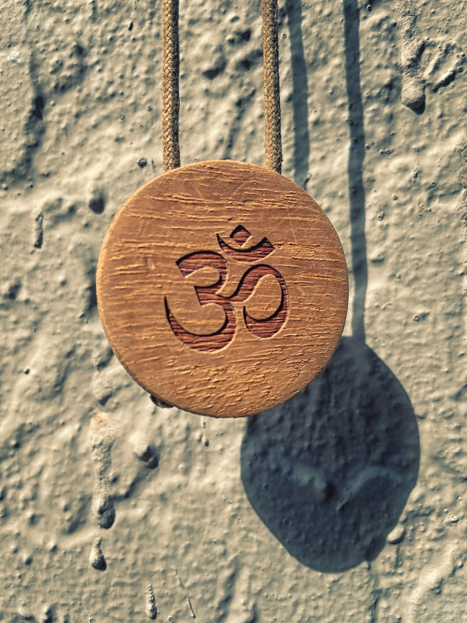 A wooden pendant with the Om symbol carved into it, hanging from a string against a textured wall.