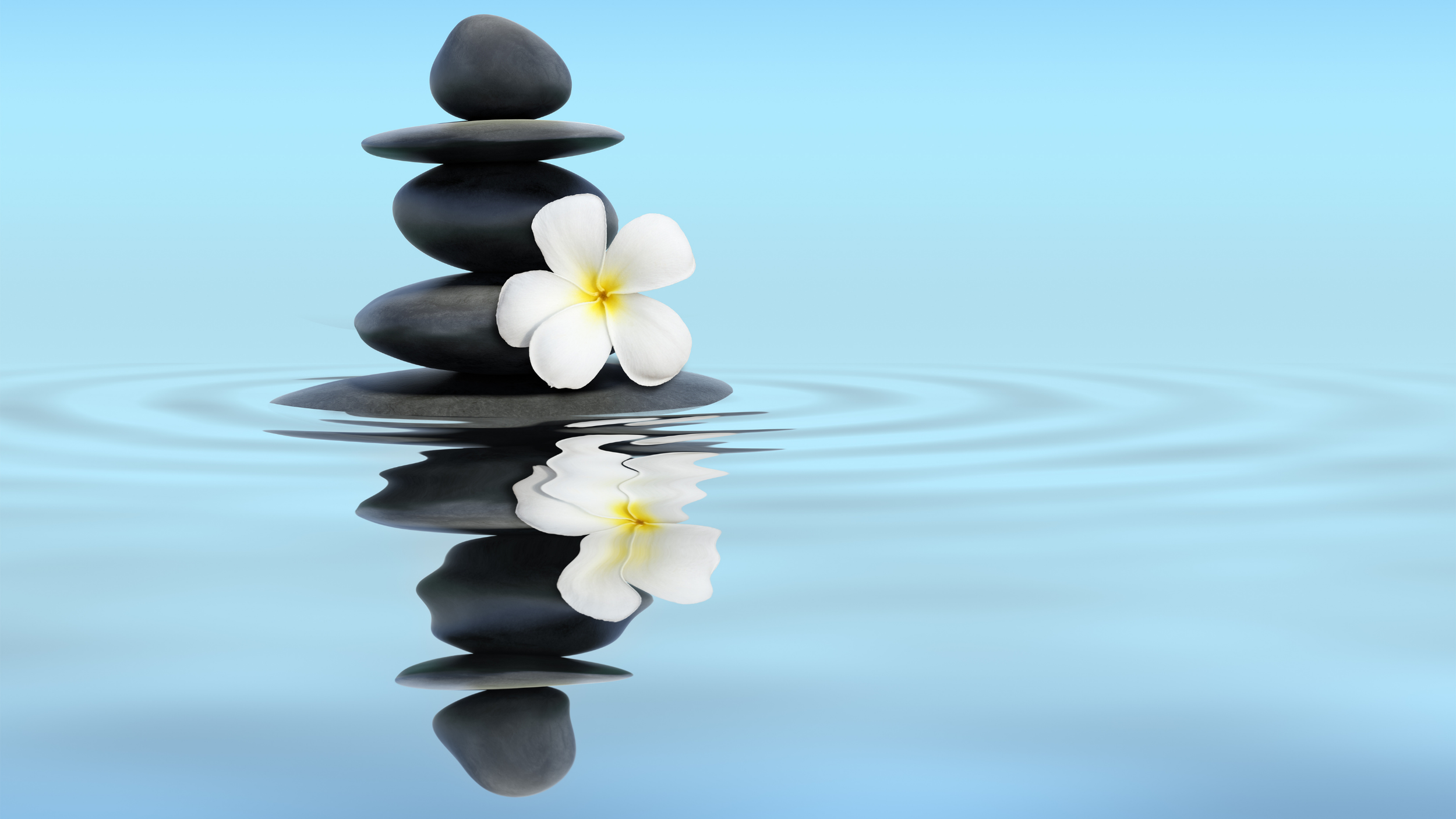 Stacked smooth black stones with a white flower beside them, reflected in calm water with a clear blue sky in the background.