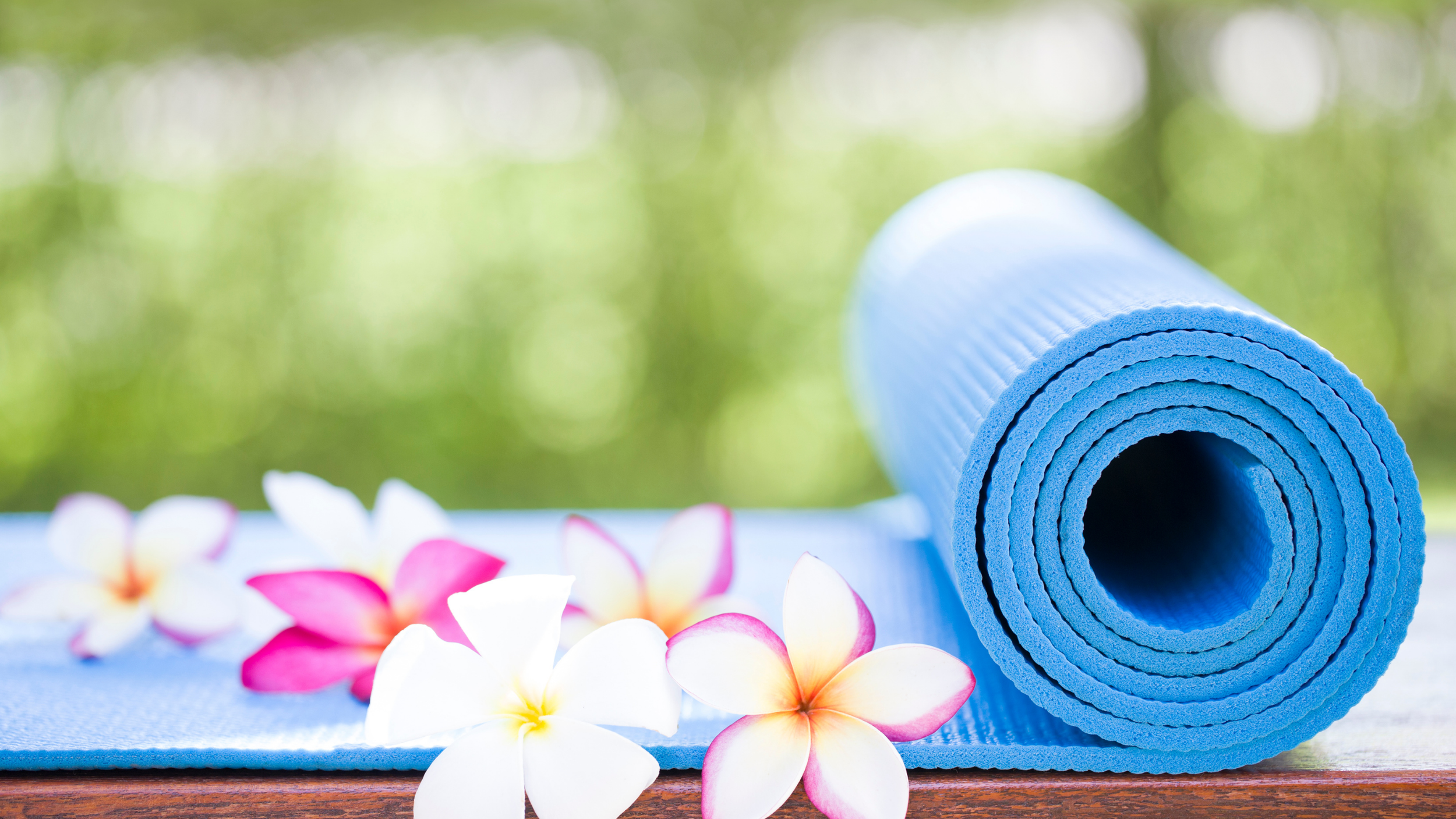 A rolled blue yoga mat on a wooden surface with white and pink flowers around it, blurred green background.