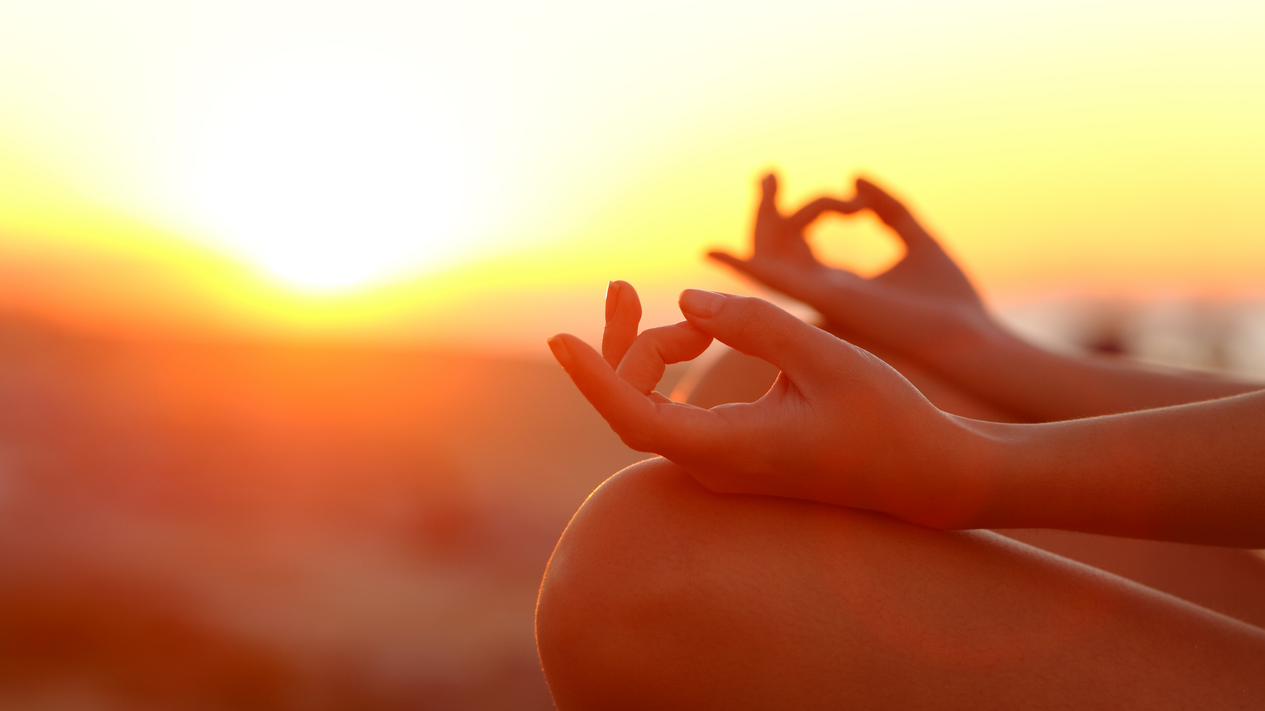 Close-up of a person's hand in a meditation pose during sunset, with another hand in the background forming a heart shape.