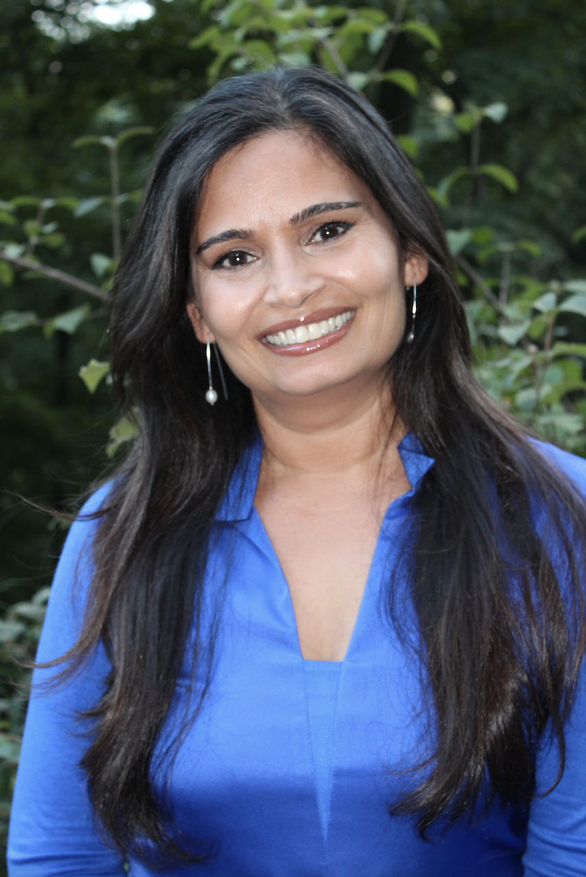 A woman with long dark hair and earrings, smiling and wearing a bright blue shirt, outdoors with green foliage in the background.