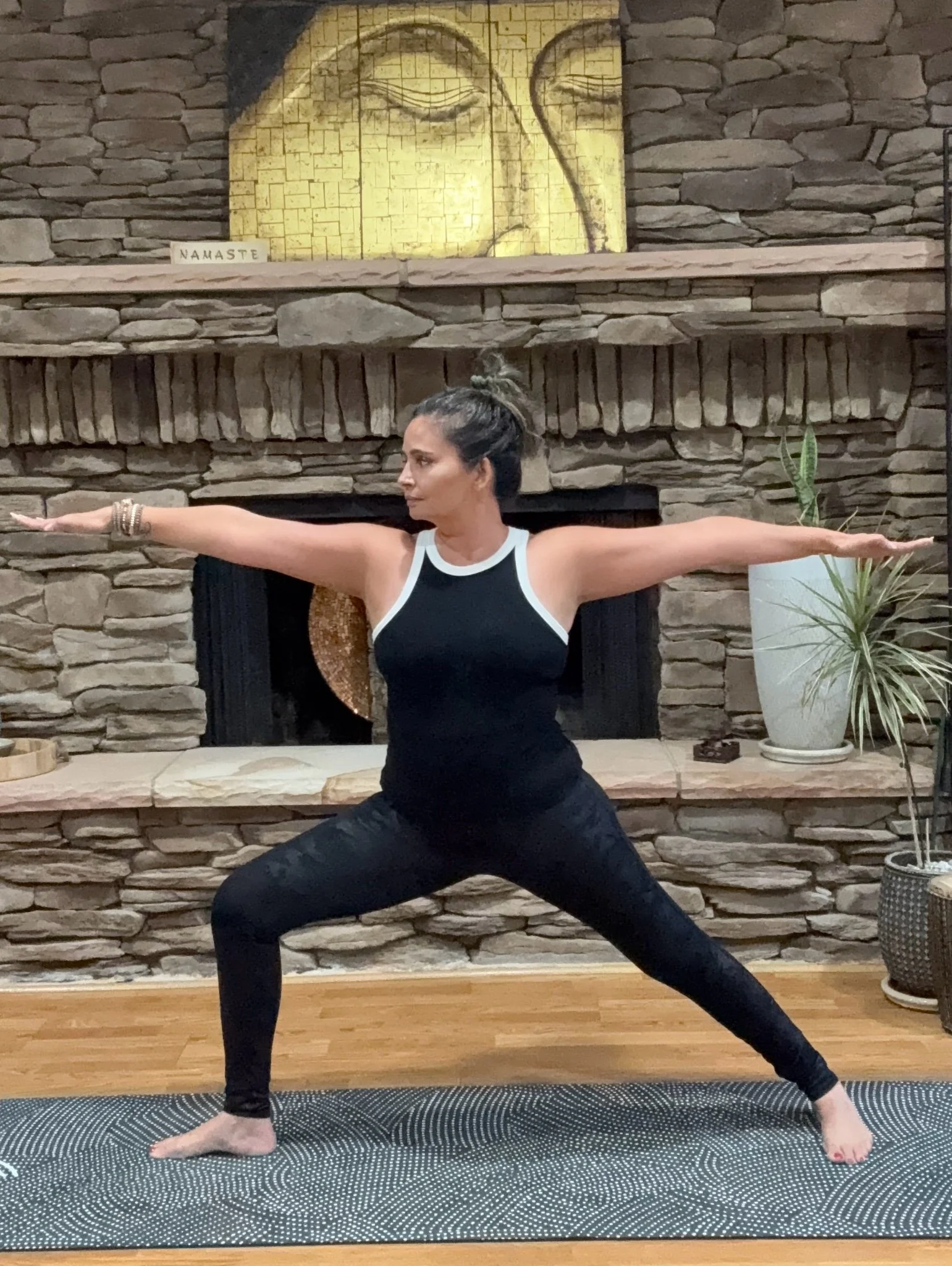 A woman practicing yoga in a room with a stone fireplace and potted plants. She is standing in a Warrior pose with arms extended, wearing a black tank top and black leggings on a yoga mat.