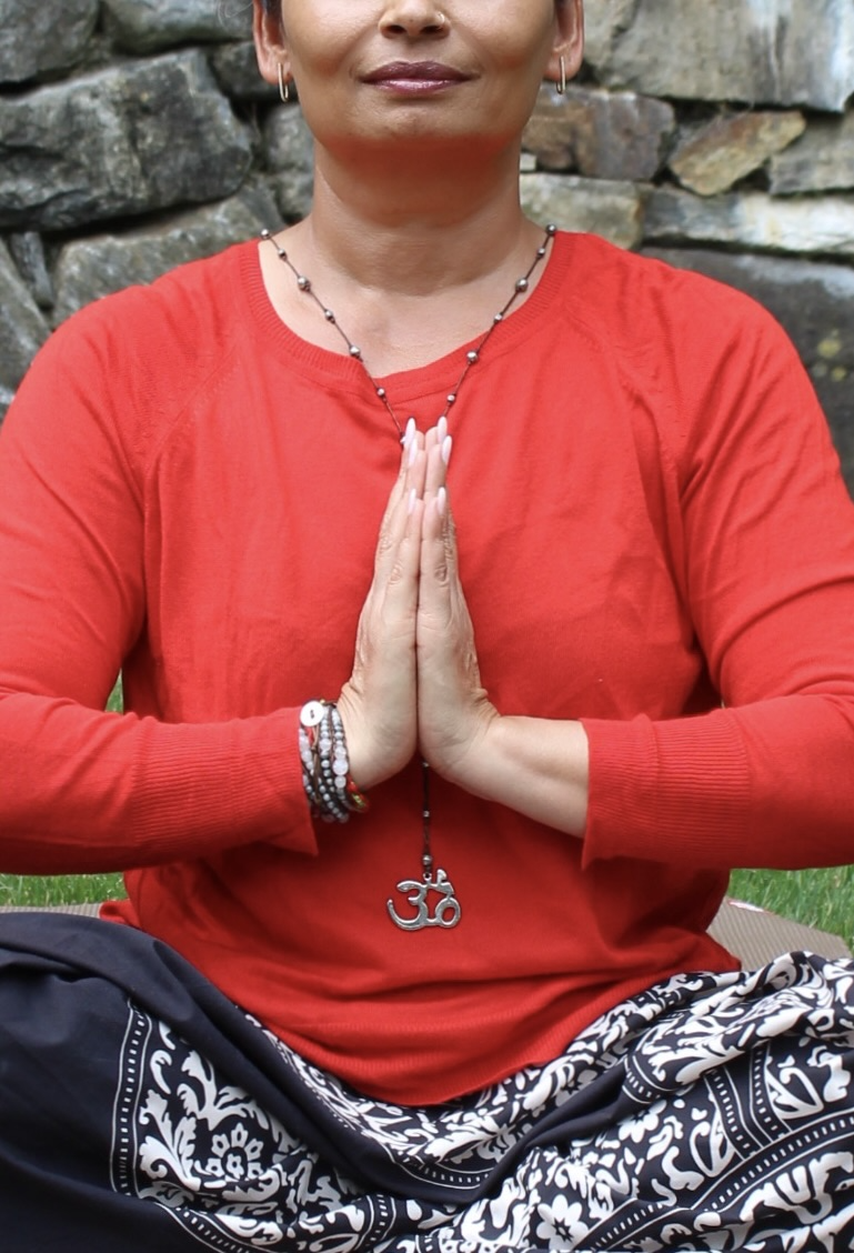 Woman practicing yoga outdoors, wearing a red top and patterned black and white pants, sitting cross-legged with hands in prayer position, jewelry including a necklace with Om symbol, bracelets, earrings, against a stone wall background.