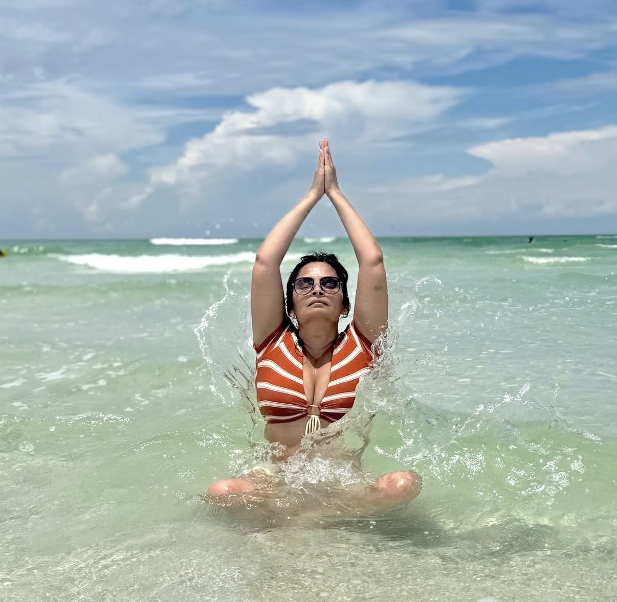 woman in sunglasses and swimsuit practicing yoga in the ocean with blue sky and clouds in background