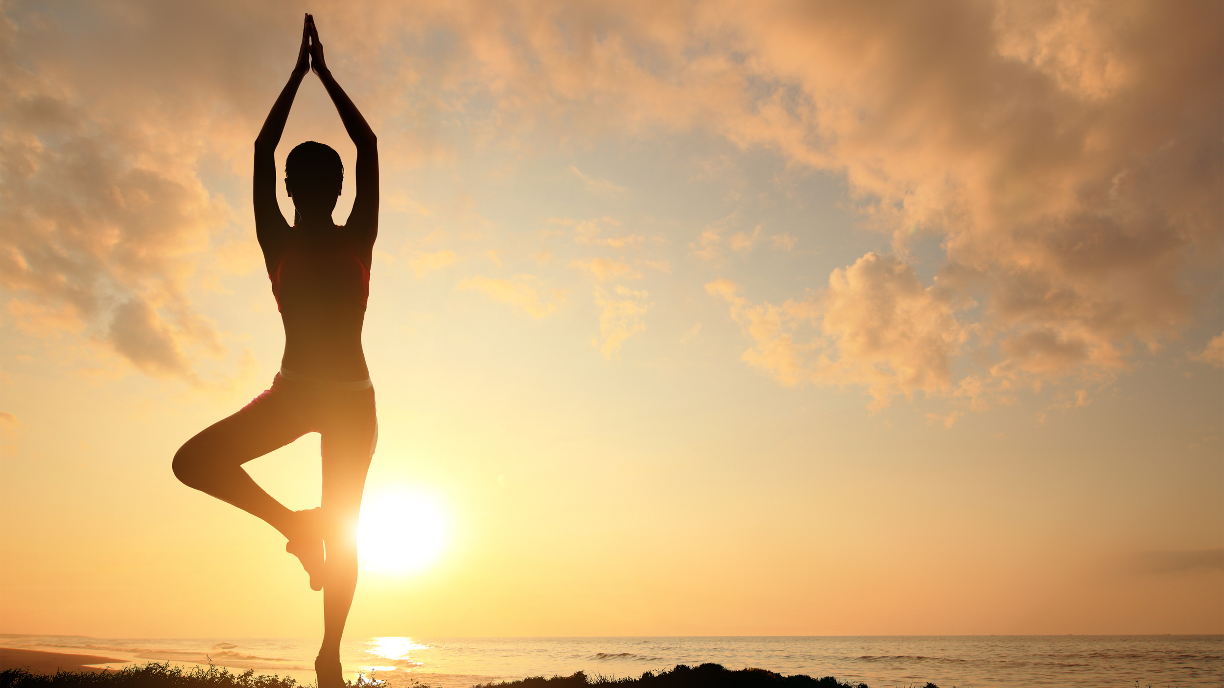 A person practicing yoga in a tree pose at sunset by the beach, with clouds in the sky.