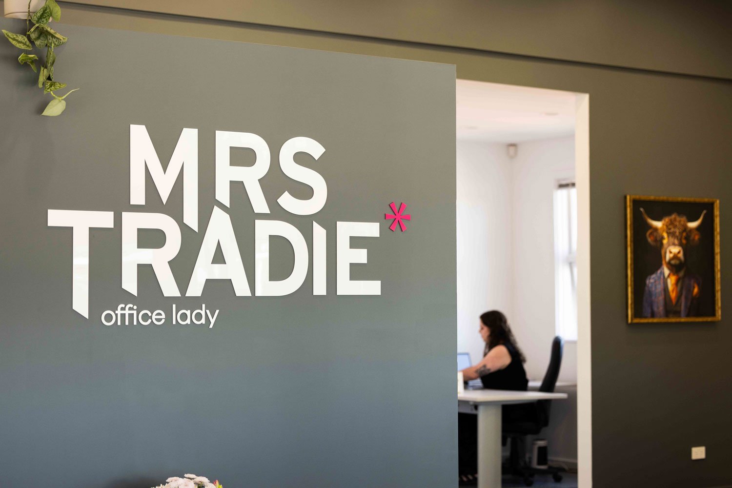 Office door with sign reading 'MRS TRADIE' and 'office lady' underneath, with a woman working at her desk visible in the background.