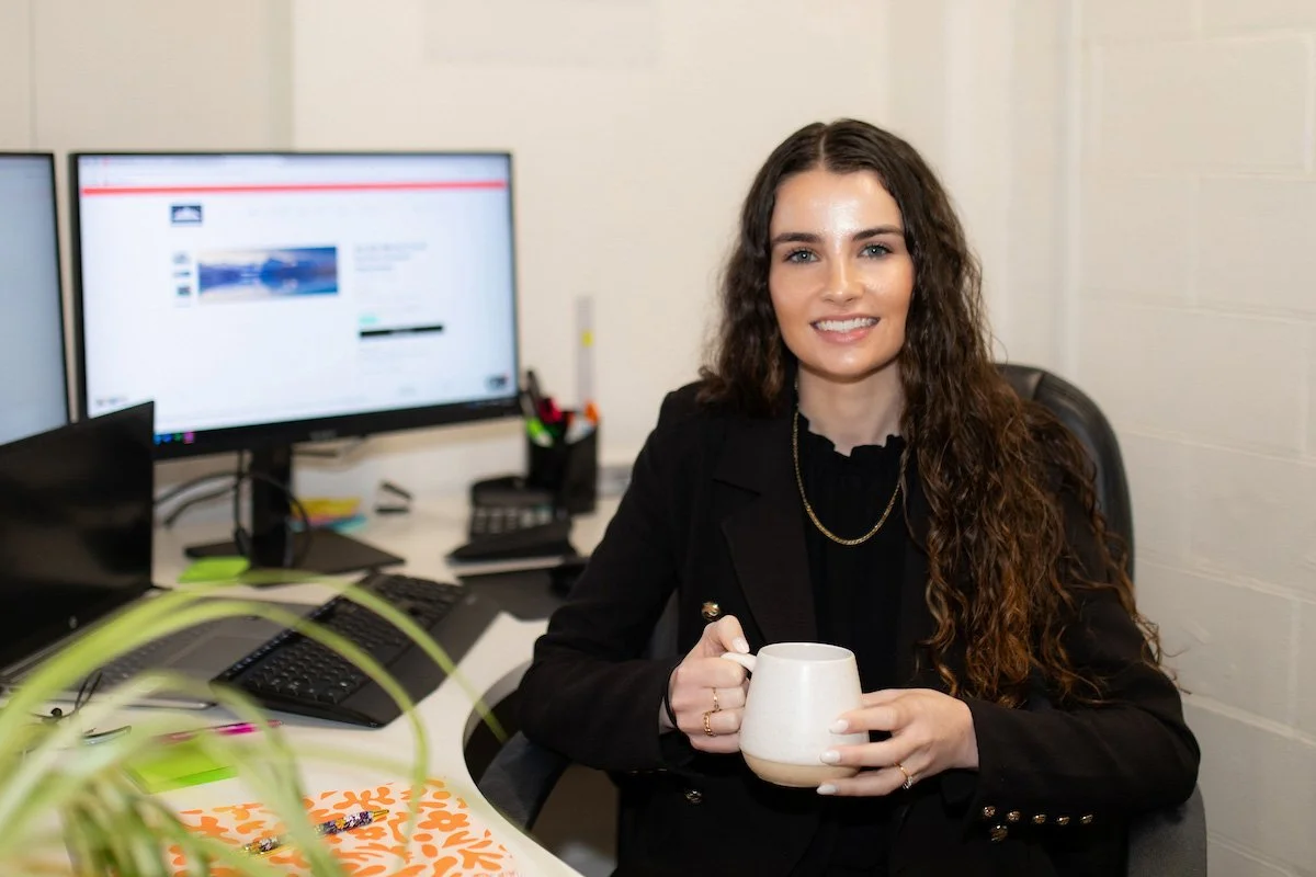 reagan sitting at mrs tradie office desk smiling, holding mug