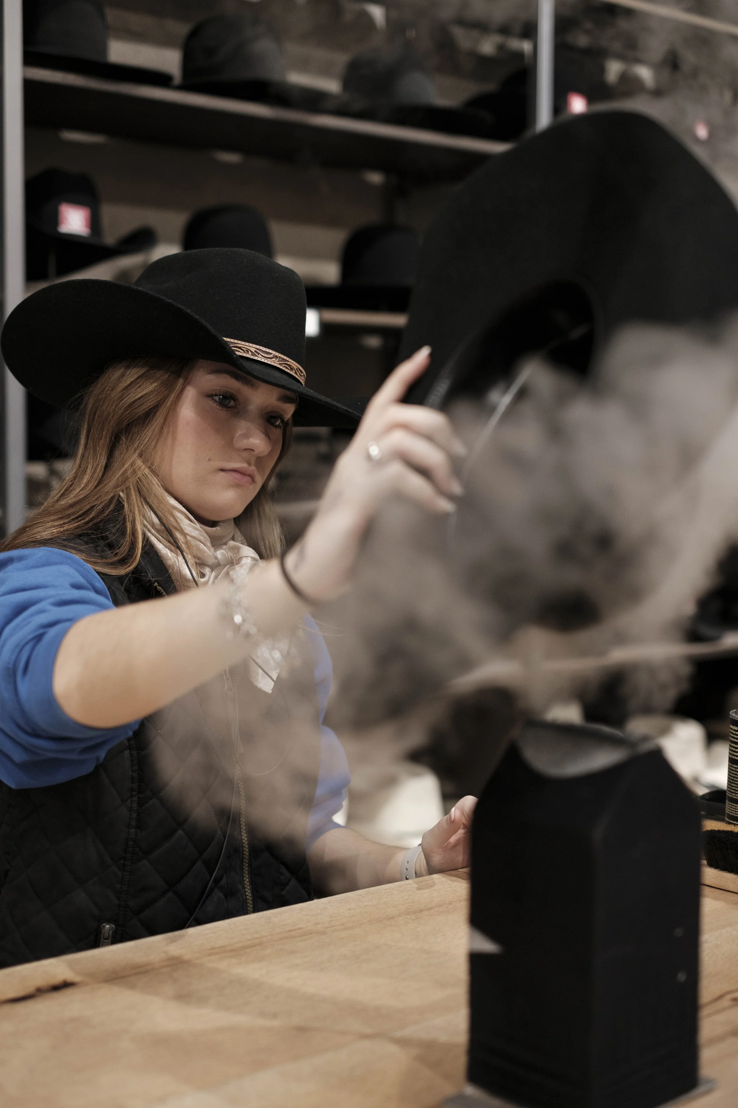 A young craftswoman shaping and brushing a black cowboy hat inside a traditional hat workshop, captured in soft indoor light, highlighting skilled hands, artisanal heritage, and the continuity of Western craftsmanship.