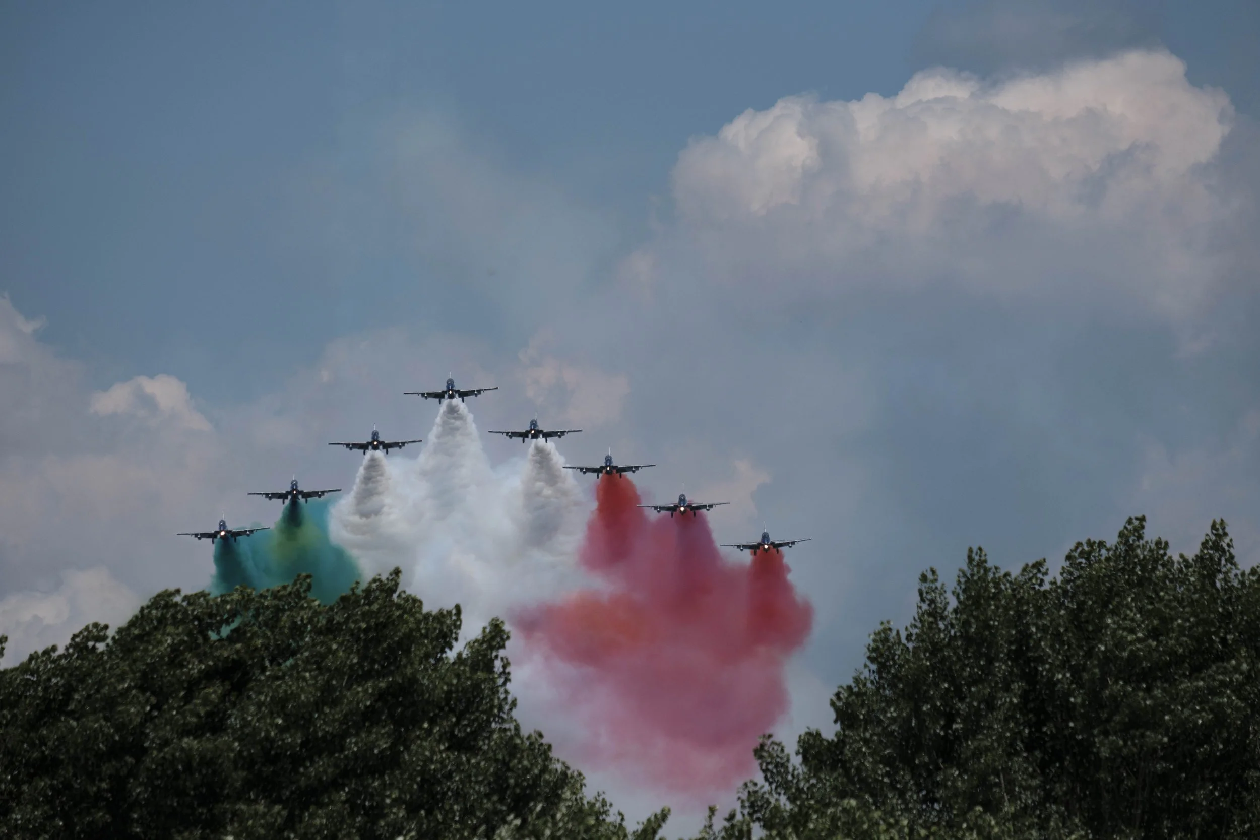 Aerobatic team flying in tight formation above treetops, releasing green, white, and red smoke trails against a partly cloudy sky, capturing precision flight, symmetry, and national colors during an air show.