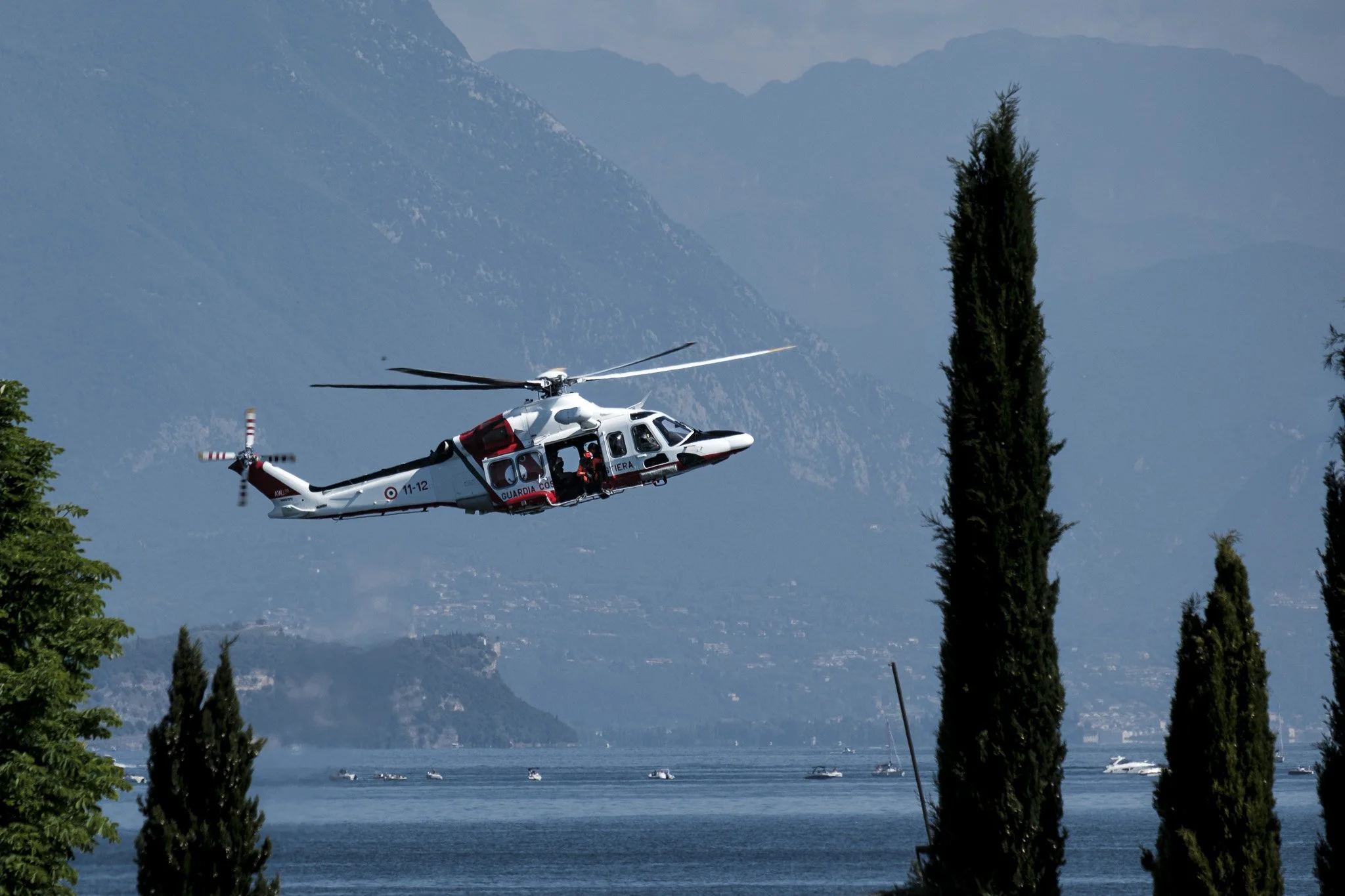 Rescue helicopter flying low over a lake with mountainous landscape in the background, framed by tall trees, capturing an aerial emergency operation in a natural alpine environment under clear daylight.