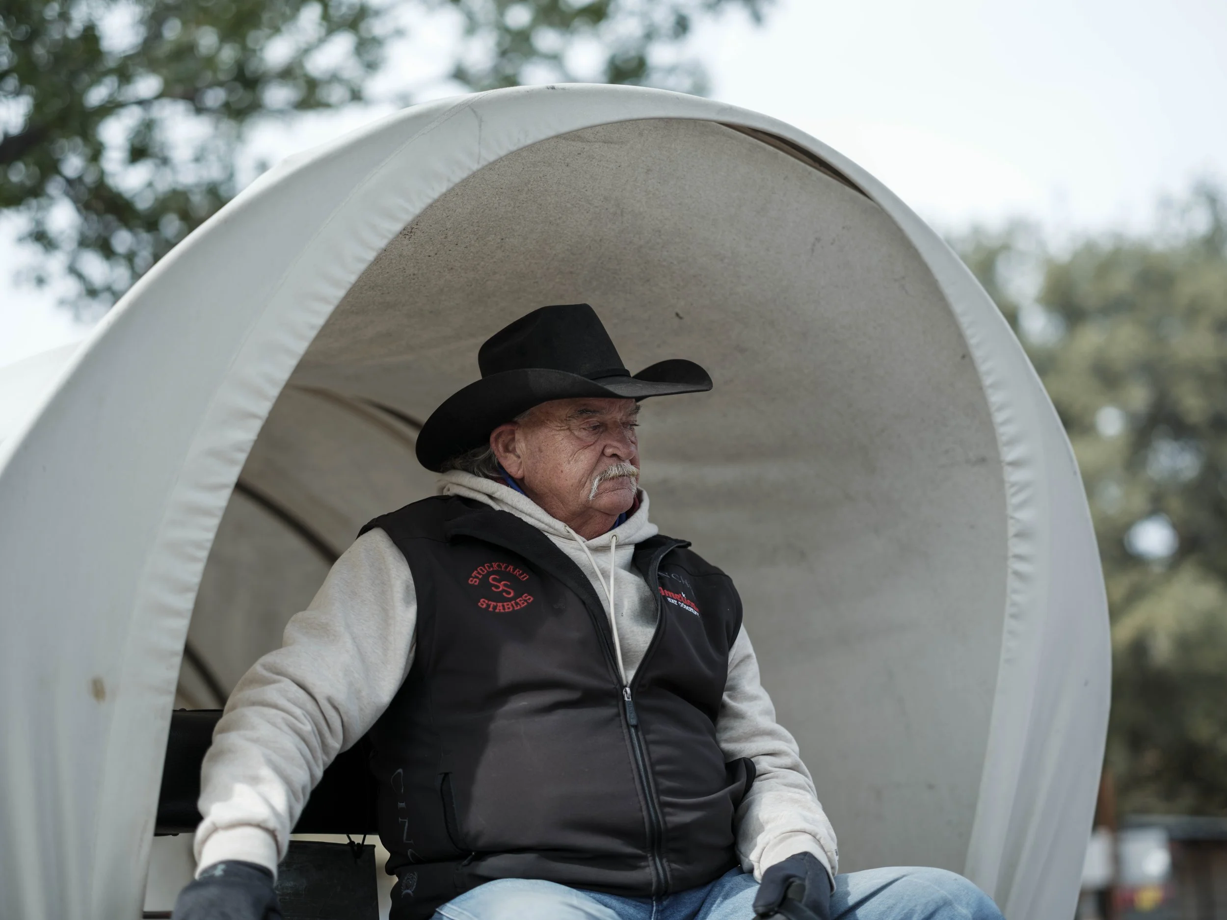 An elderly cowboy sitting beneath a covered wagon canopy, wearing a black hat and layered work clothing, photographed in soft natural daylight, conveying a sense of frontier heritage, quiet resilience, and timeless rural tradition.
