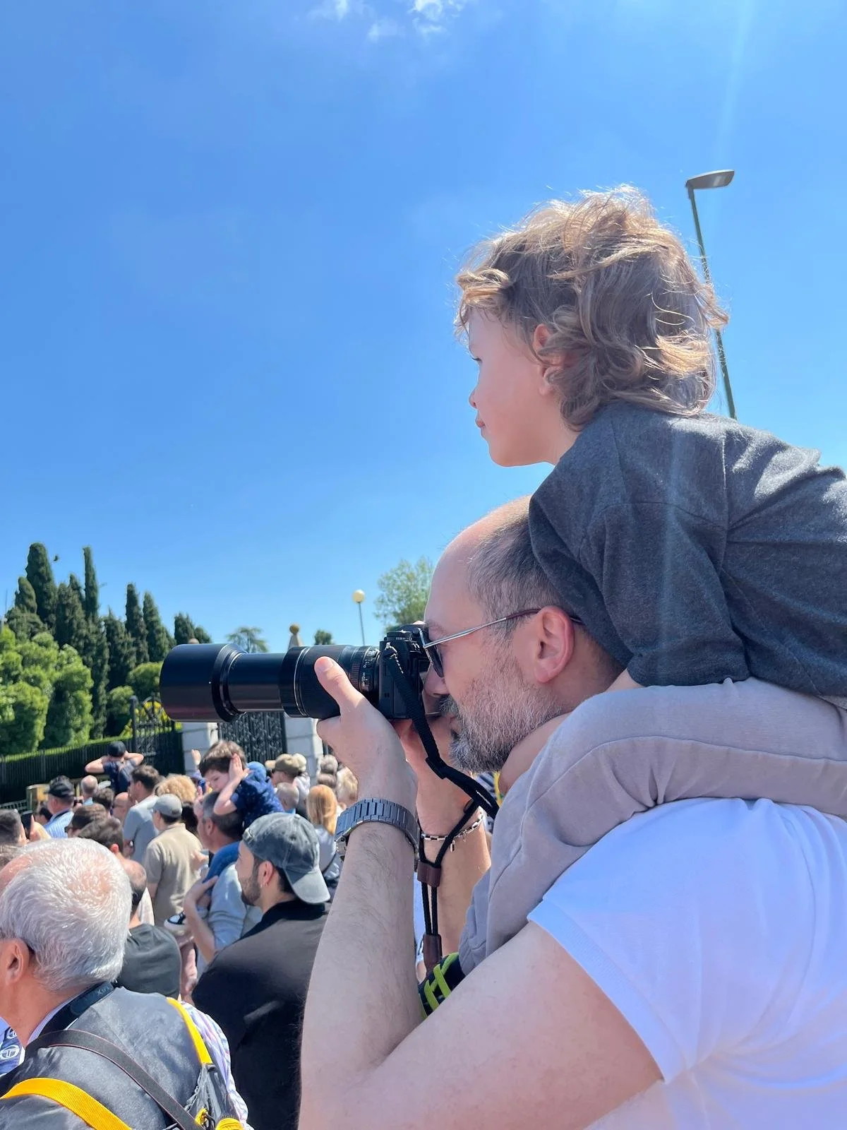 A photographer observing a scene with a child on his shoulders, capturing a moment of quiet attention within a crowded environment.