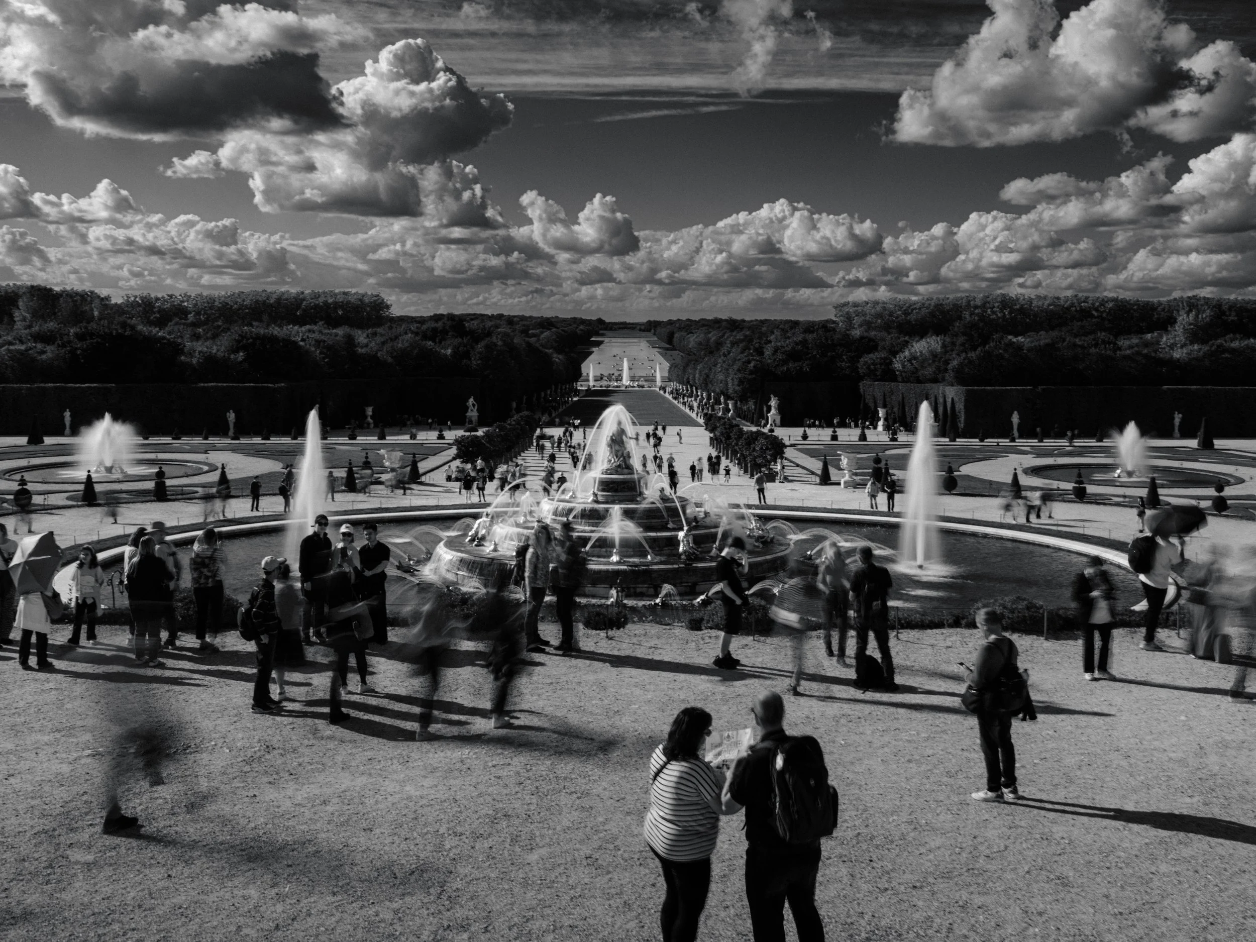 Crowded formal garden with a central fountain and long reflecting axis, captured in black and white, showing blurred visitors in motion beneath dramatic clouds, emphasizing symmetry, scale, and the contrast between architecture and human presence.