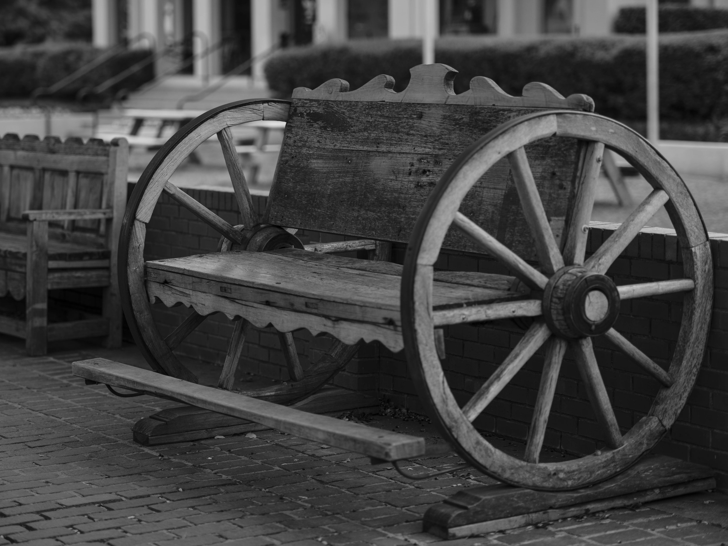 An old wooden cart bench with large wagon wheels displayed in an outdoor public space, photographed in black and white with soft natural light, highlighting weathered textures, rural craftsmanship, and a sense of stillness within an urban setting.