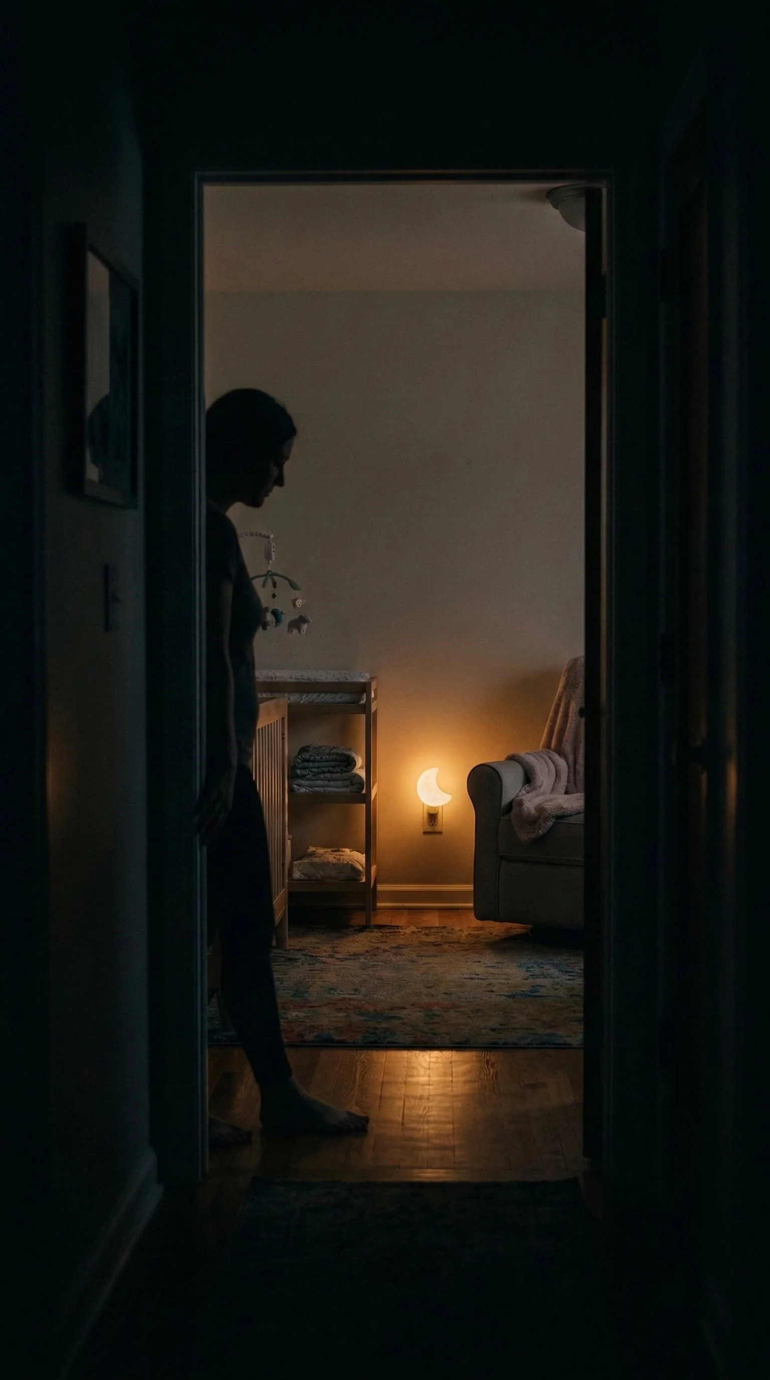 A woman stands in silhouette within a dark doorway, looking into a quiet nursery. The room is softly lit by a small, moon shaped nightlight plugged into the wall, casting an orange glow over a rocking chair and a crib.