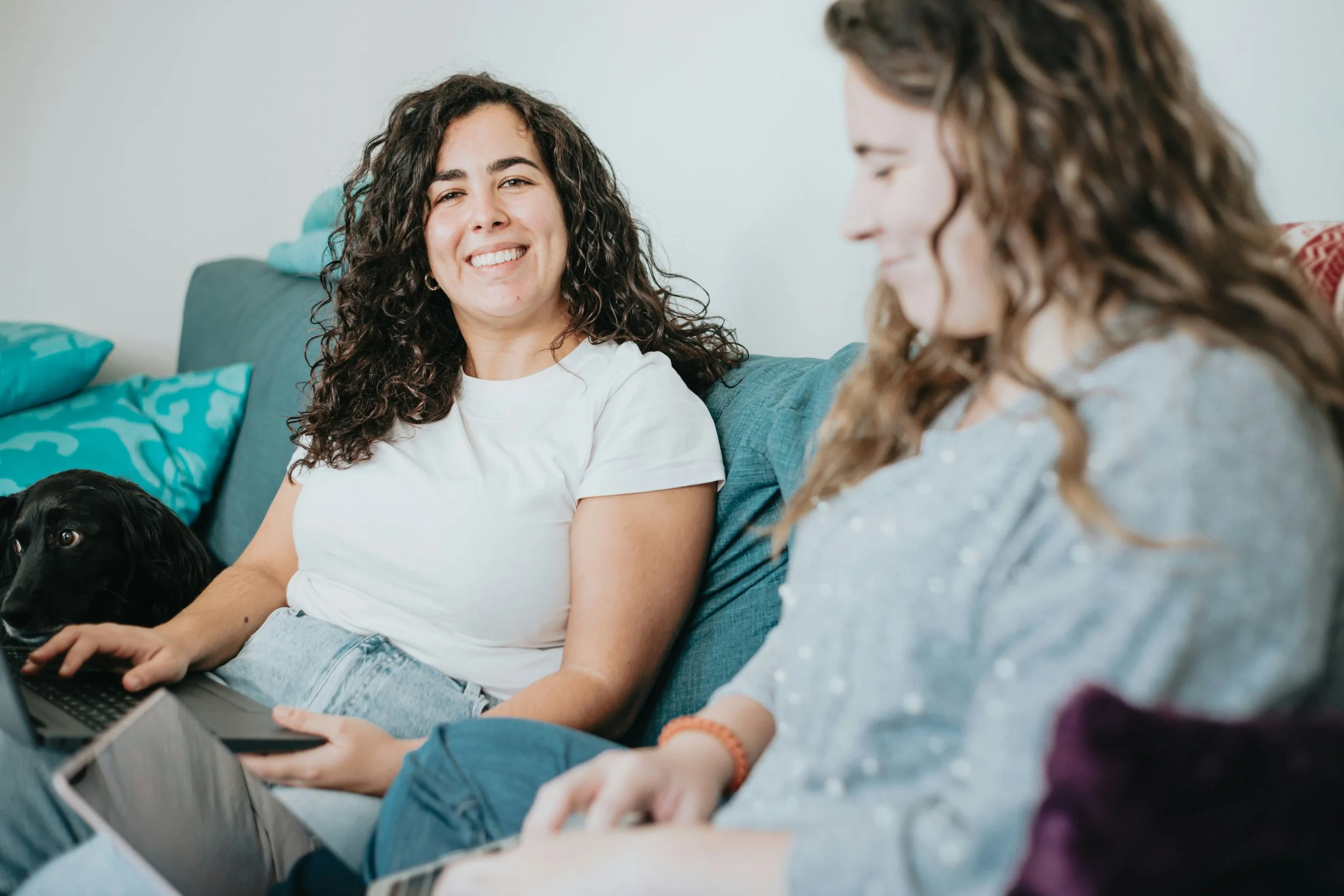 Two women laughing together on a couch, with a black dog tucked beside them