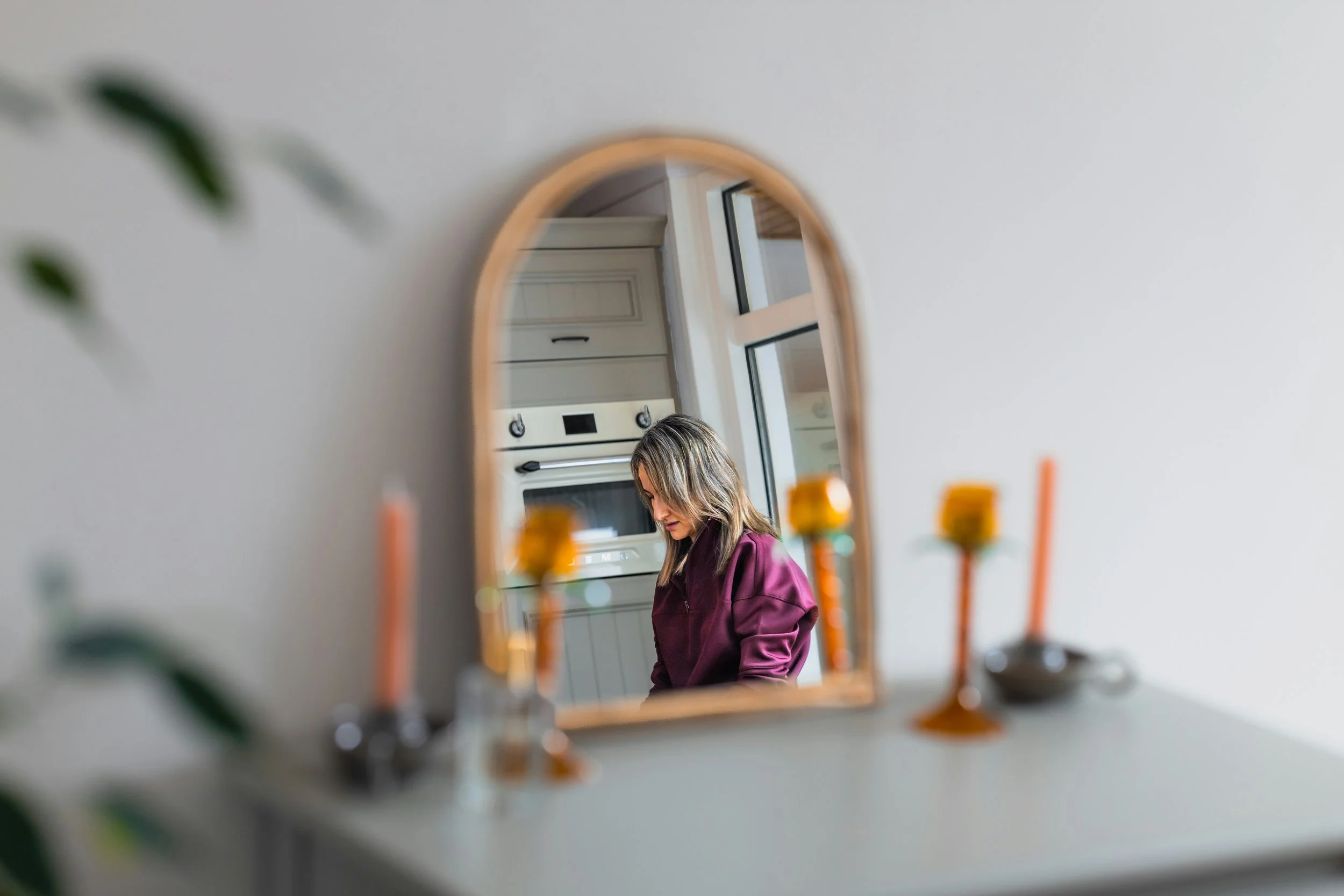 A woman reflected in an arched mirror in her kitchen, looking downward in a quiet moment of thought